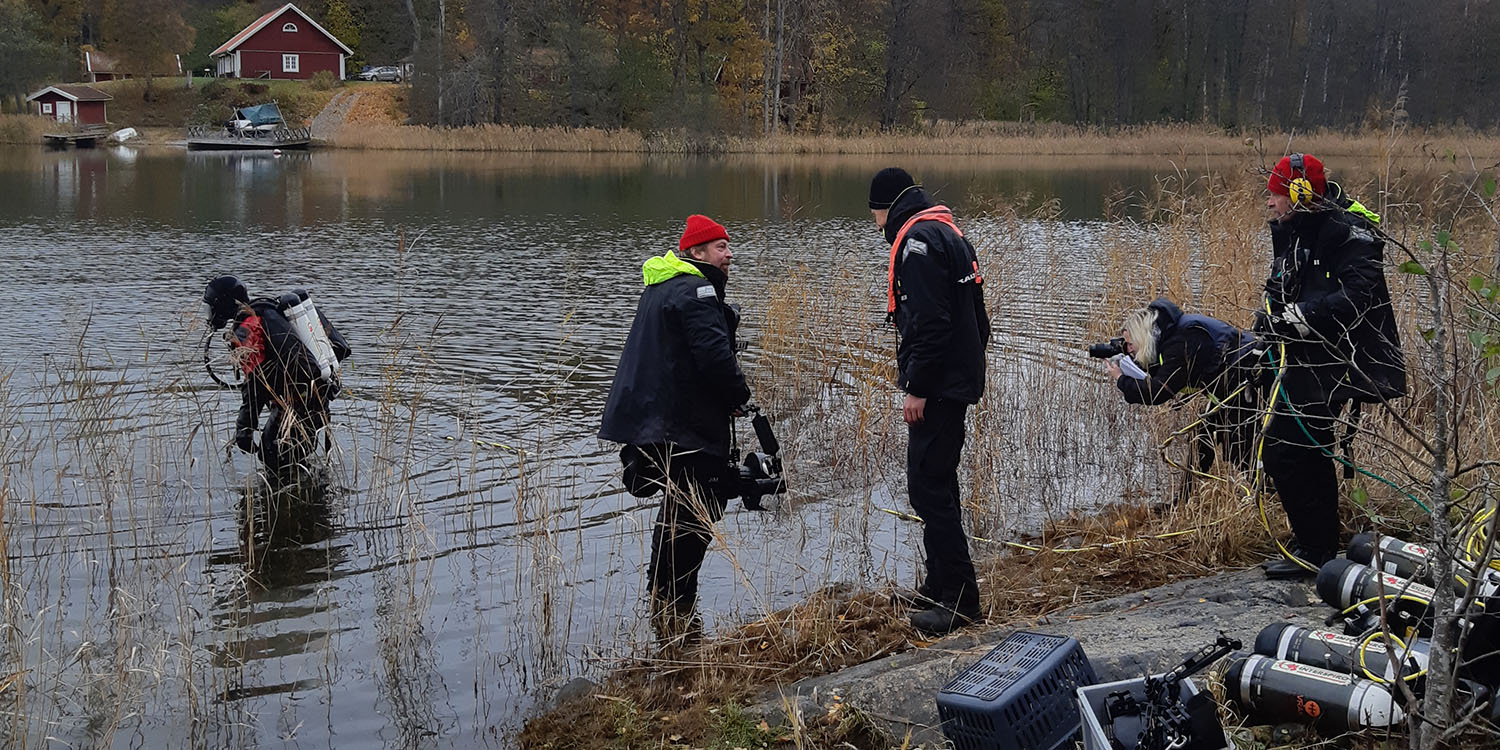 Några personer med dykarutrustning och fotoutrustning står vid vattenbrynet medan en dykare är på väg ner i vattnet. 