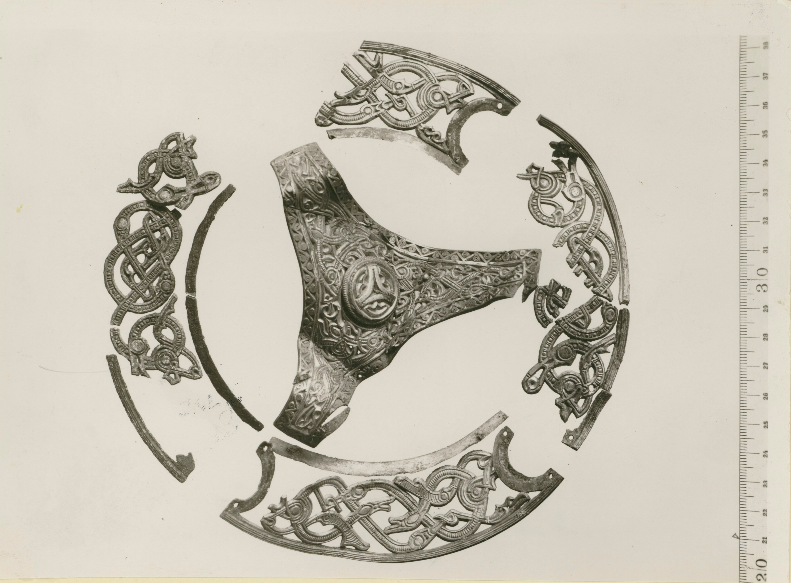 Flera fragment av ett rikt ornamenterat metallföremål, arrangerade i cirkelform på ljus bakgrund. En linjal syns längs bildens högra kant.