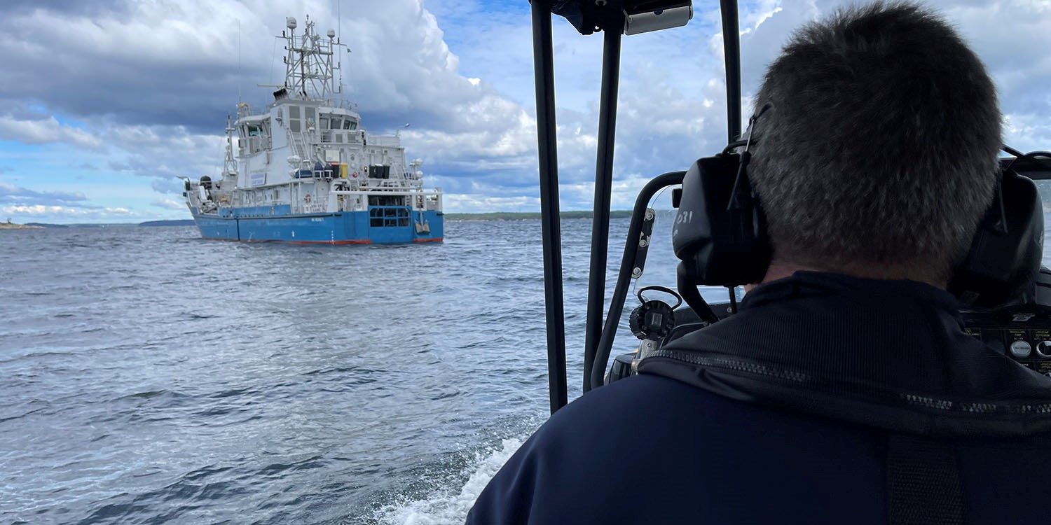 En man sedd bakifrån styrandes en båt i riktning mot ett annat fartyg. 