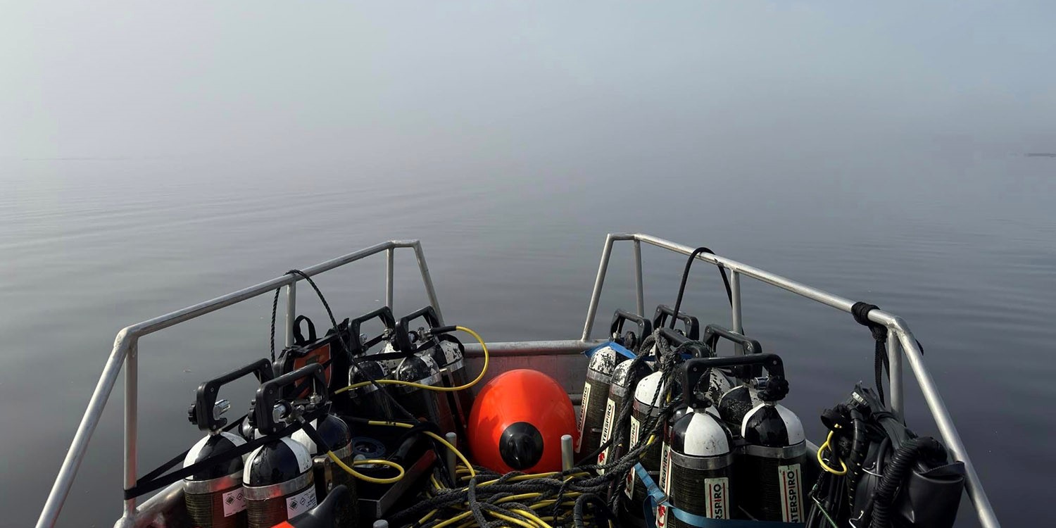 Fören på en båt med flera dykflaskor och utrustning fastspända längs relingen samt en orange boj i mitten.