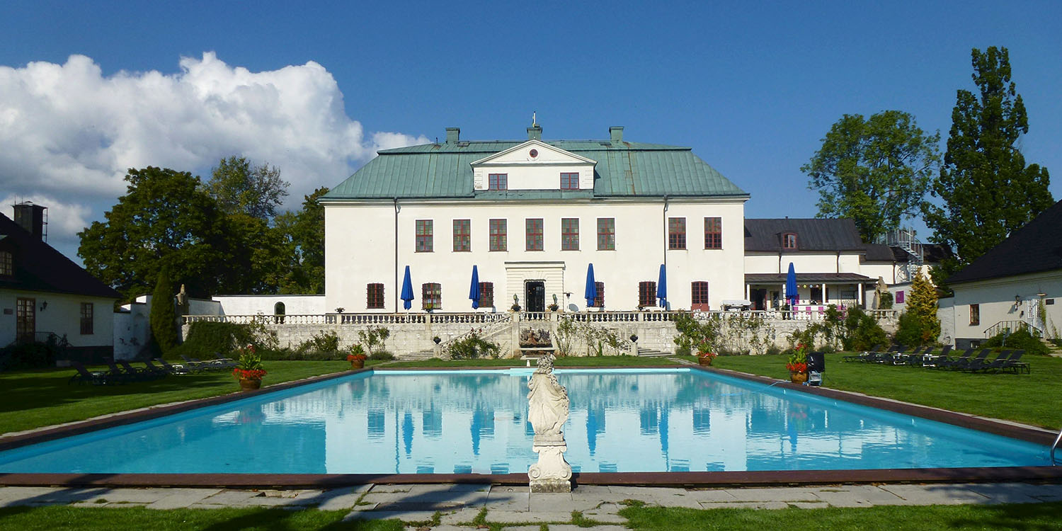 En vitrappad pampig byggnad med röda spröjs på fönstren och en stor vattenfylld pool framför.