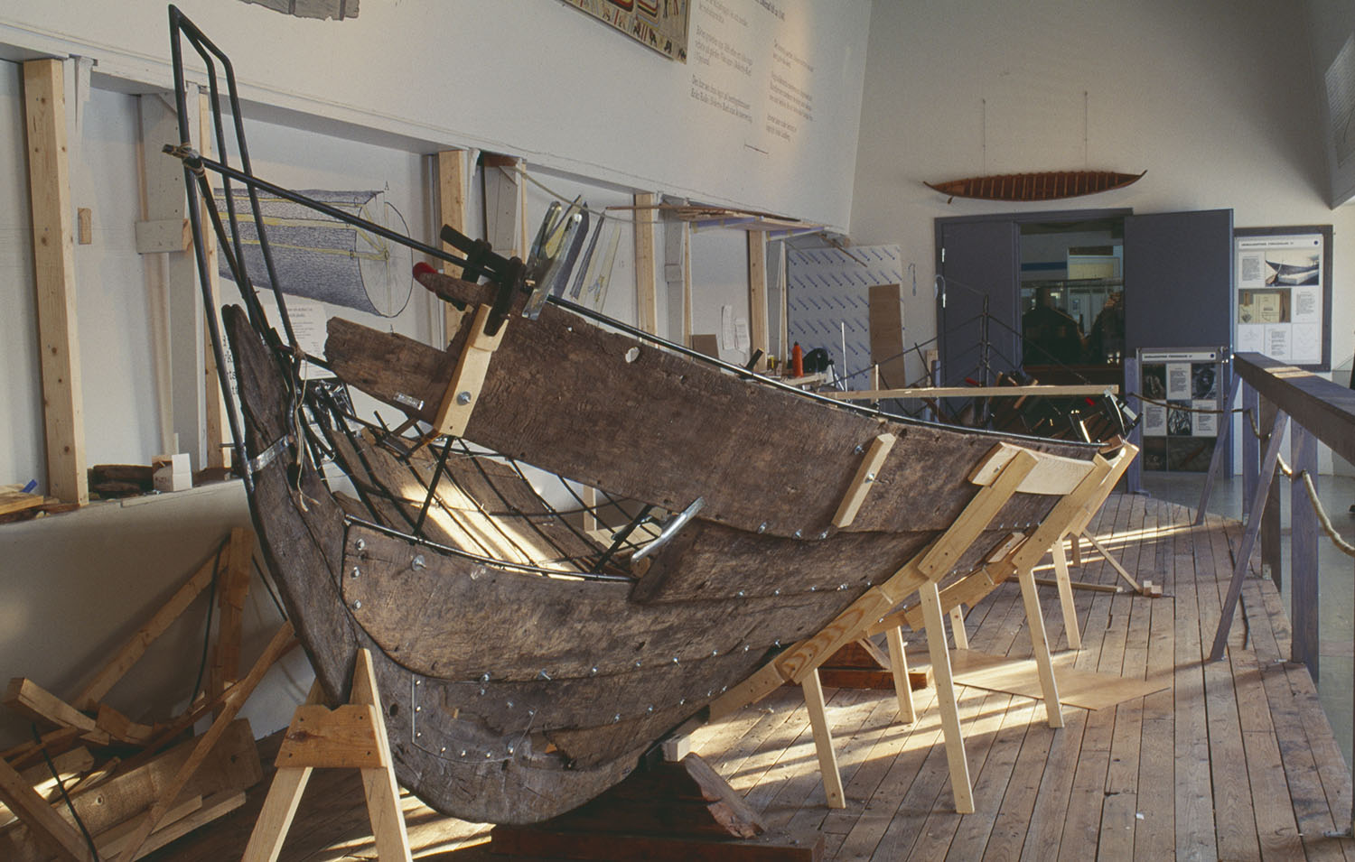 En gammal träbåt står uppställd på trästöd inomhus i en utställningslokal. 