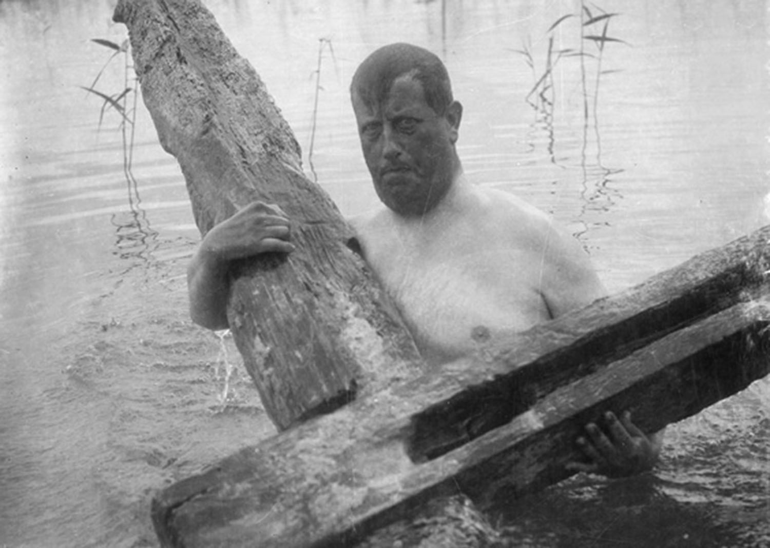 Svartvit bild av en person med bar bringa som står i vattnet med två rejäla stockar.