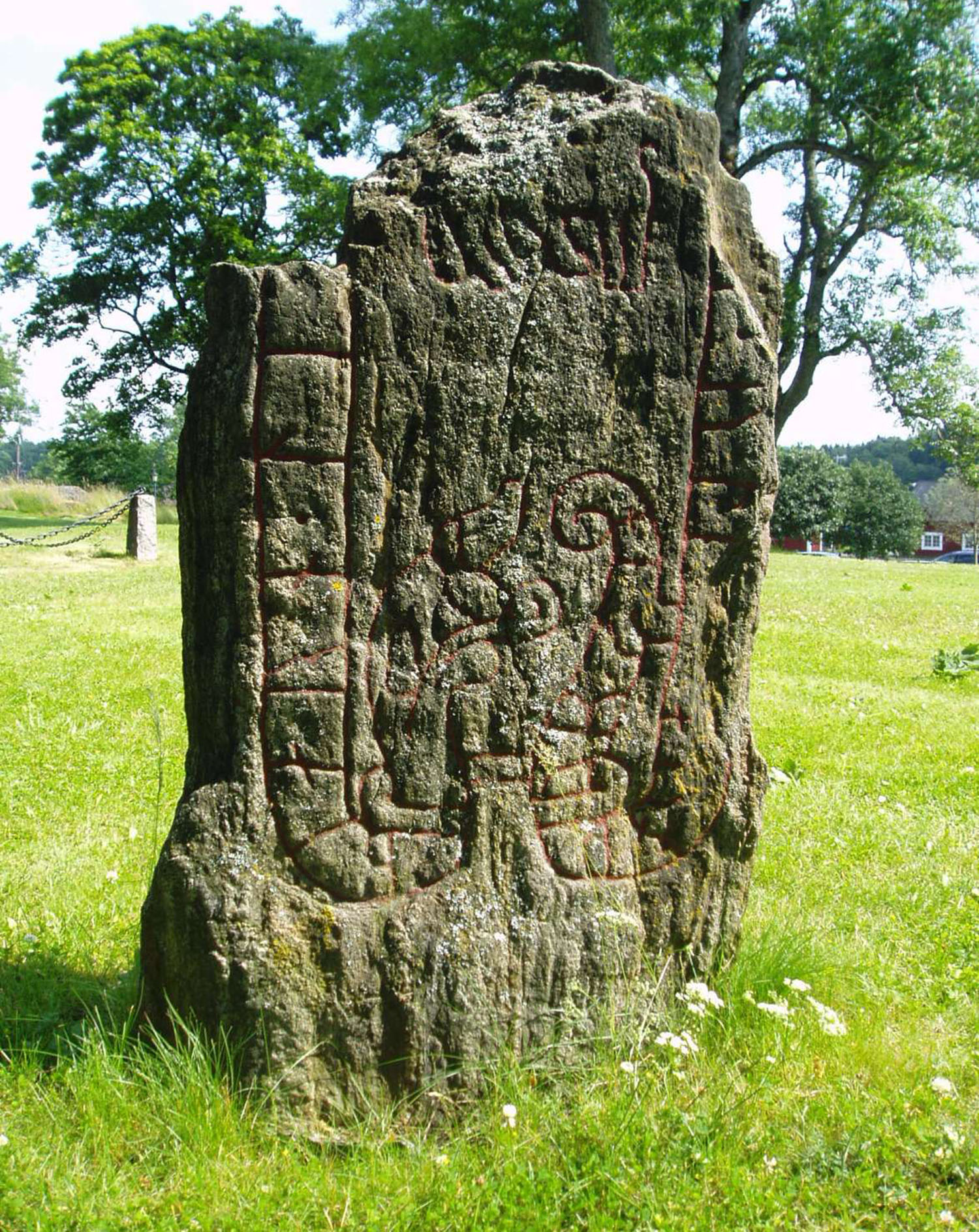 En runsten står på en äng, i bakgrunden skymtas en väg och röda hus med vita knutar.