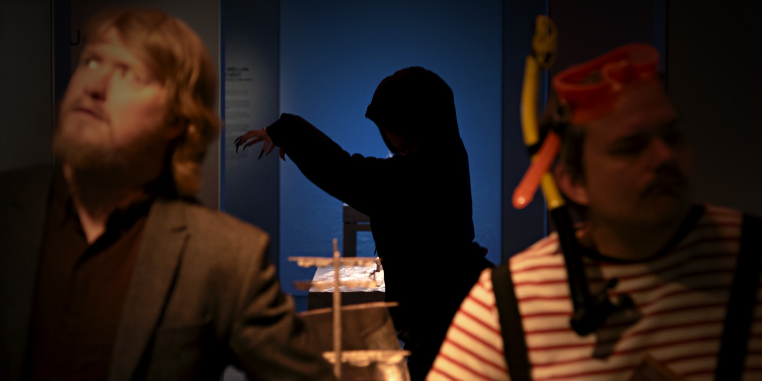 Foto av museums utställningshall i dämpad belysning. I förgrunden två personer som tittar åt olika håll, den ena har cyklop. I bakgrunden syns figur i mörk kåpa och utsträckta händer. 