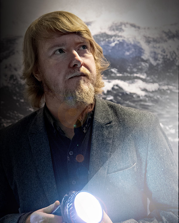 Man i mörk kavaj står framför svartvit fotovägg med havsmotiv. Han tittar åt sidan och lyser med en ficklampa. 