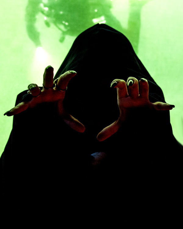 Gestalt i mörk kåpa stäcker sina händer med långa mörka klor mot betraktaren, hen syns mot grön vägg med undervattensmotiv. 