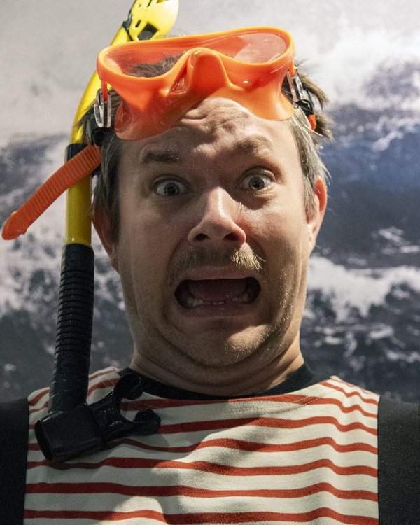 Ung man med cyklop och snorkel på huvudet ser rädd ut. I bakgrunden syns fototapet med havsmiljö. 