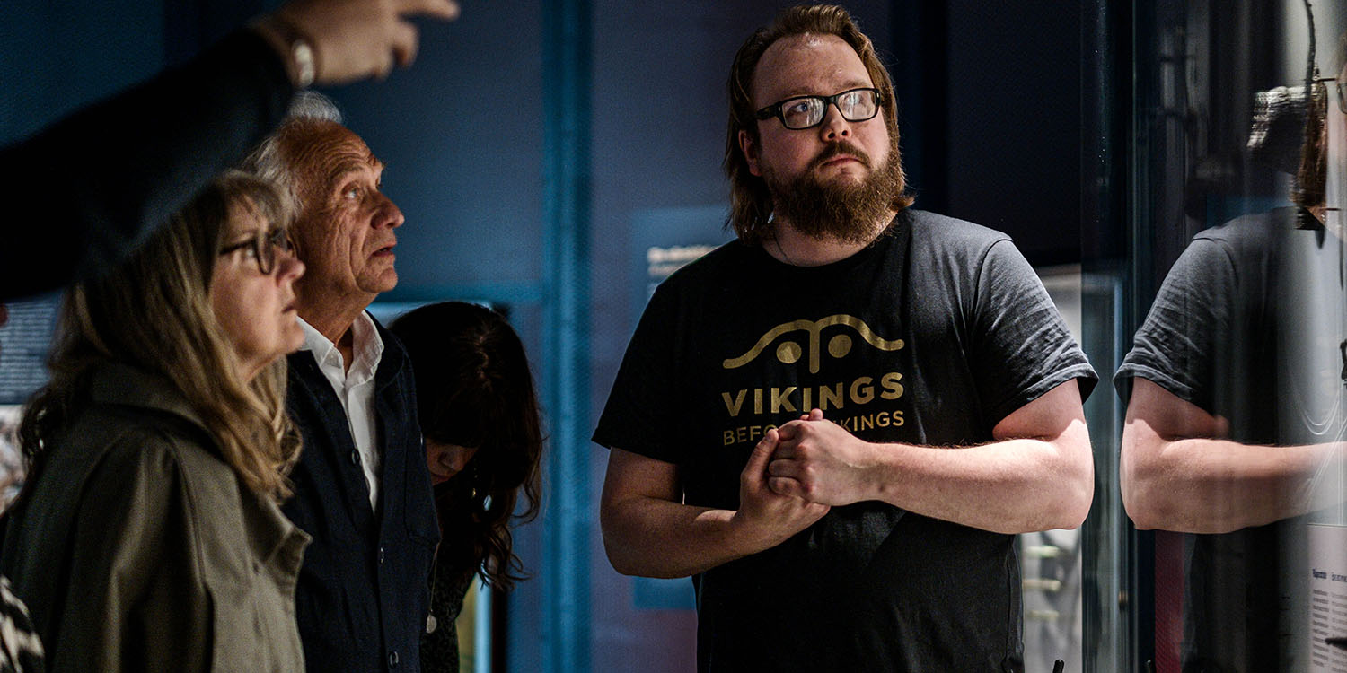 Två äldre personer i blått rum i museum, med mörkhårig guide i tröja med texten "Vikings before Vikings"