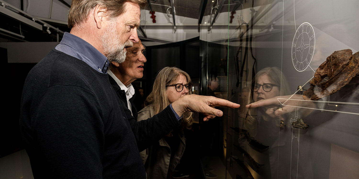 Tre äldre personer vid vägg med utställningsföremål.