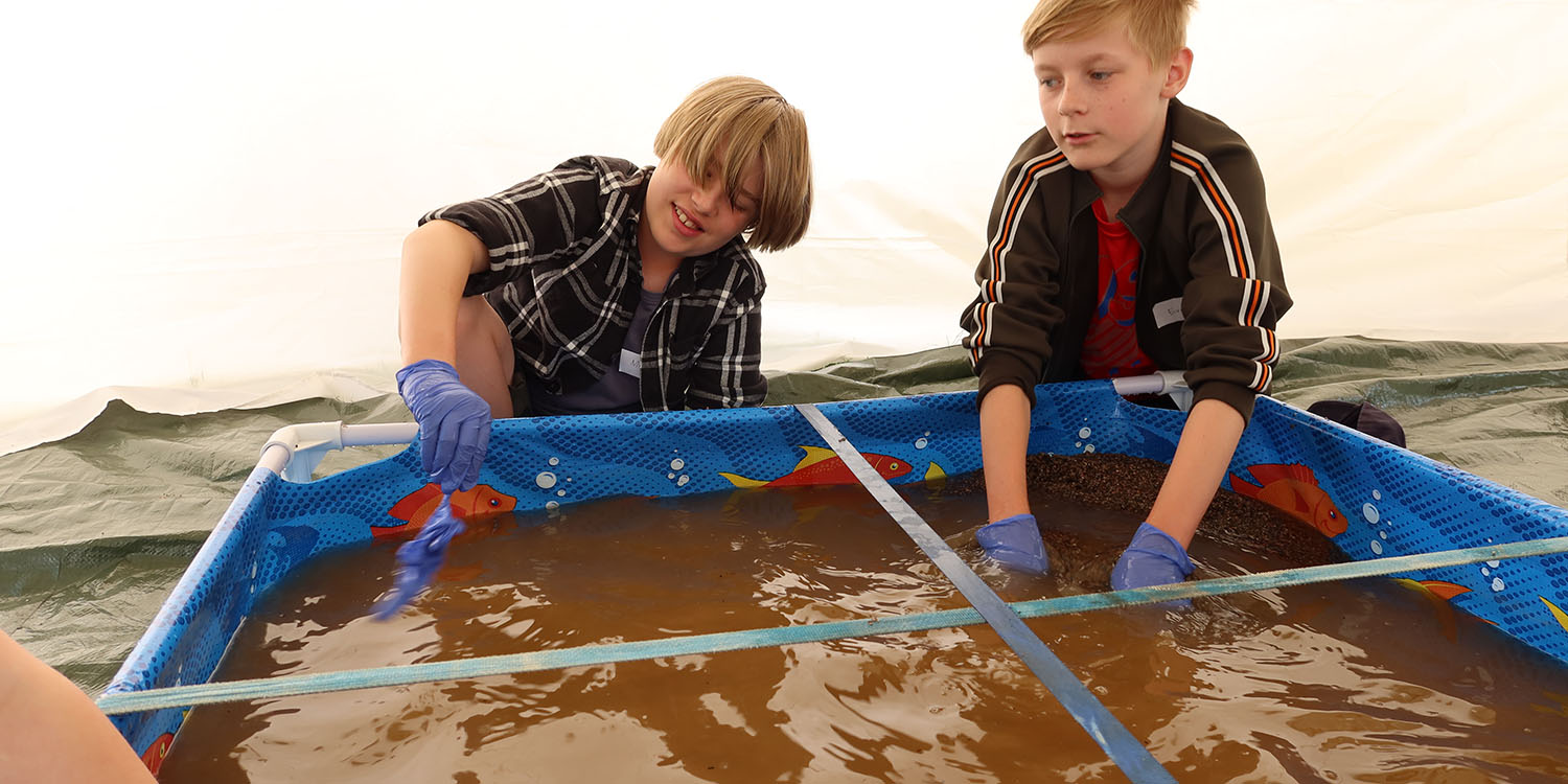 Två barn undersöker ett kar med brunfärgat vatten. De befinner sig i ett vitt tält. 