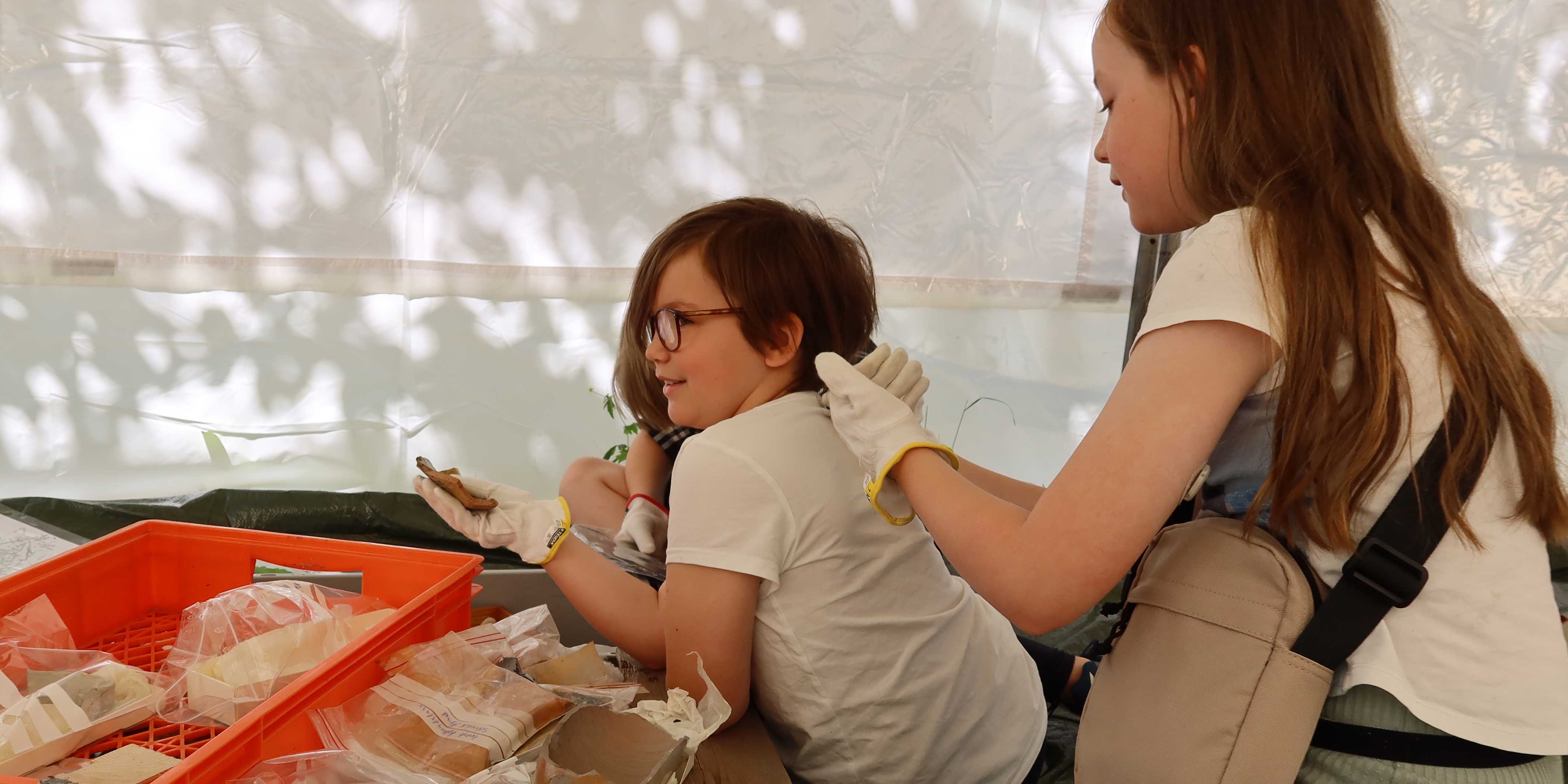 Två barn inne i vitt tält med askar med fornfynd på golvet. 