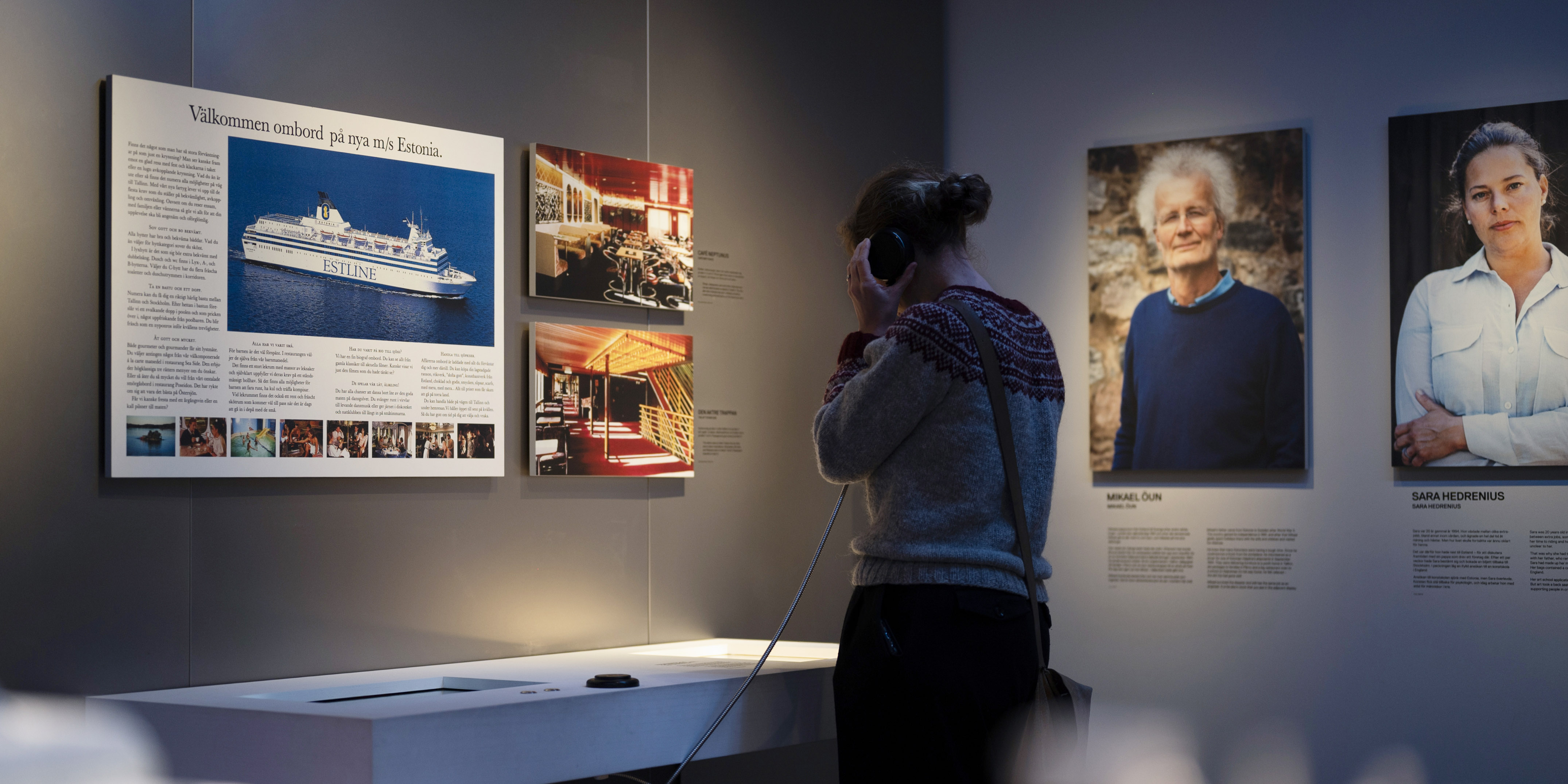 Besökare lyssnar i hörlurar framför informationsvägg om passagerarfartyget Estonia med fotografier och porträtt.