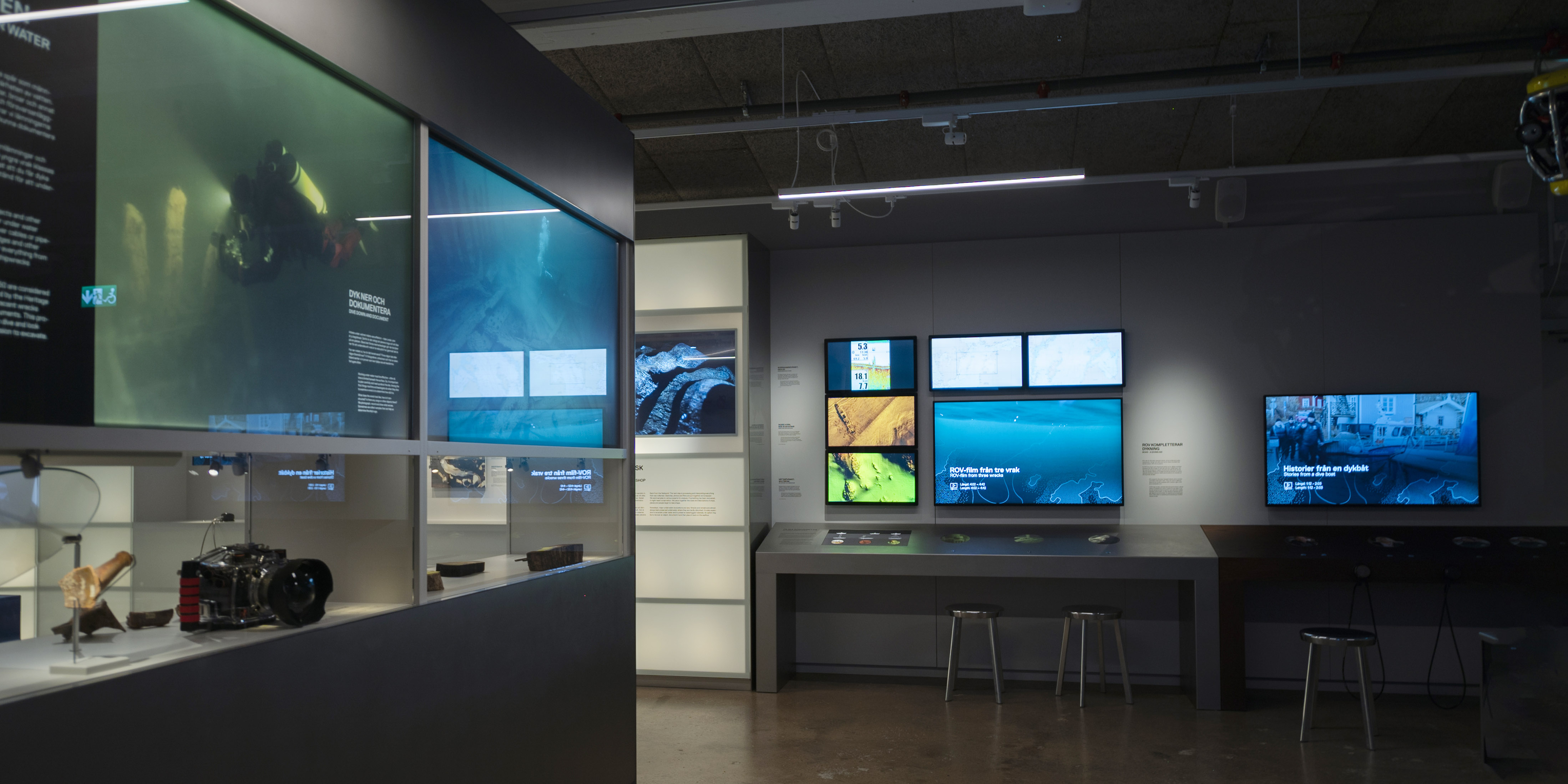 Utställning med dykutrustning i monter och flera skärmar som visar undervattensbilder och kartor.