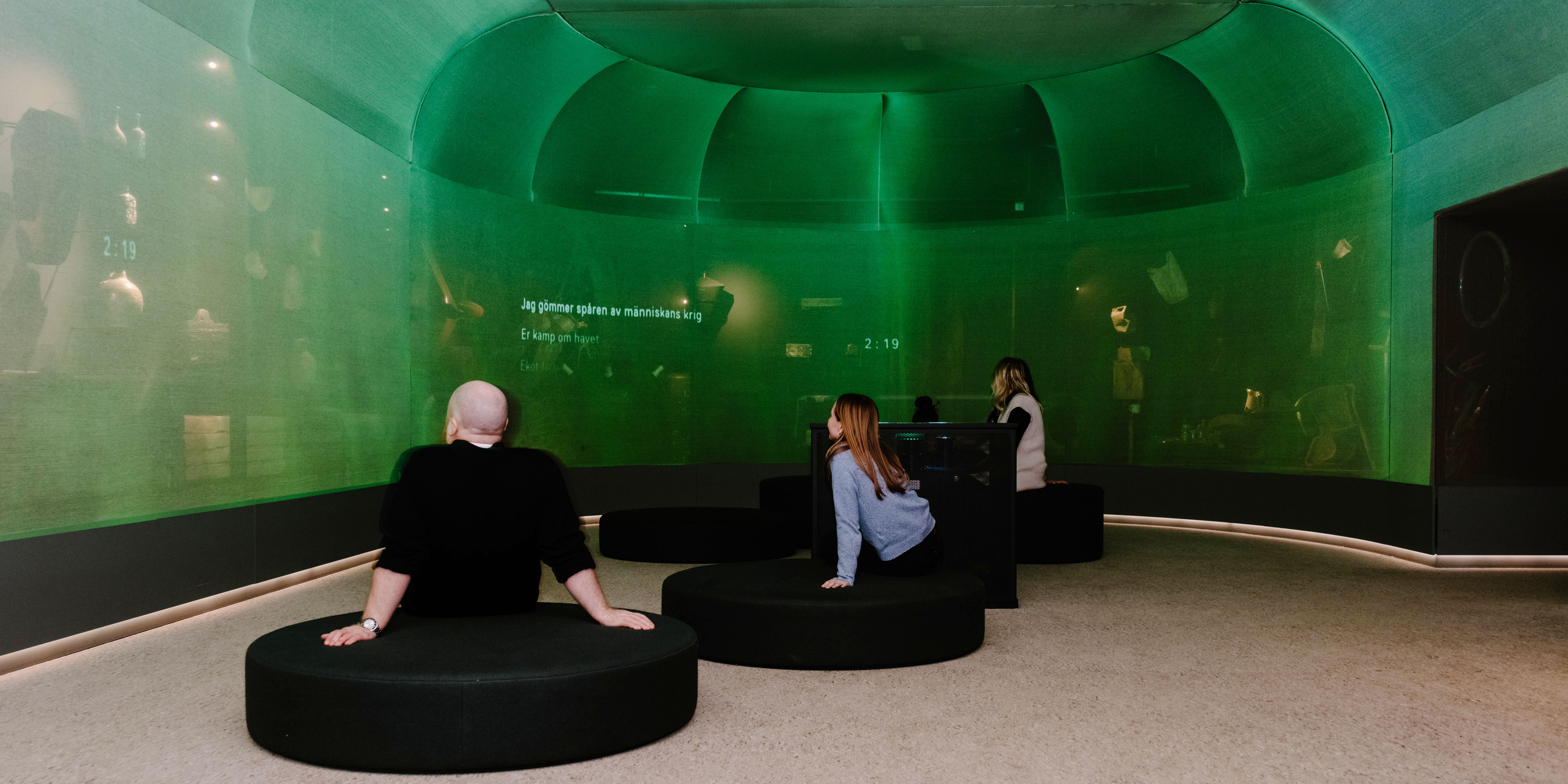 Besökare sitter i ett runt, grönt projektionrum med film och text projicerad på väggarna.