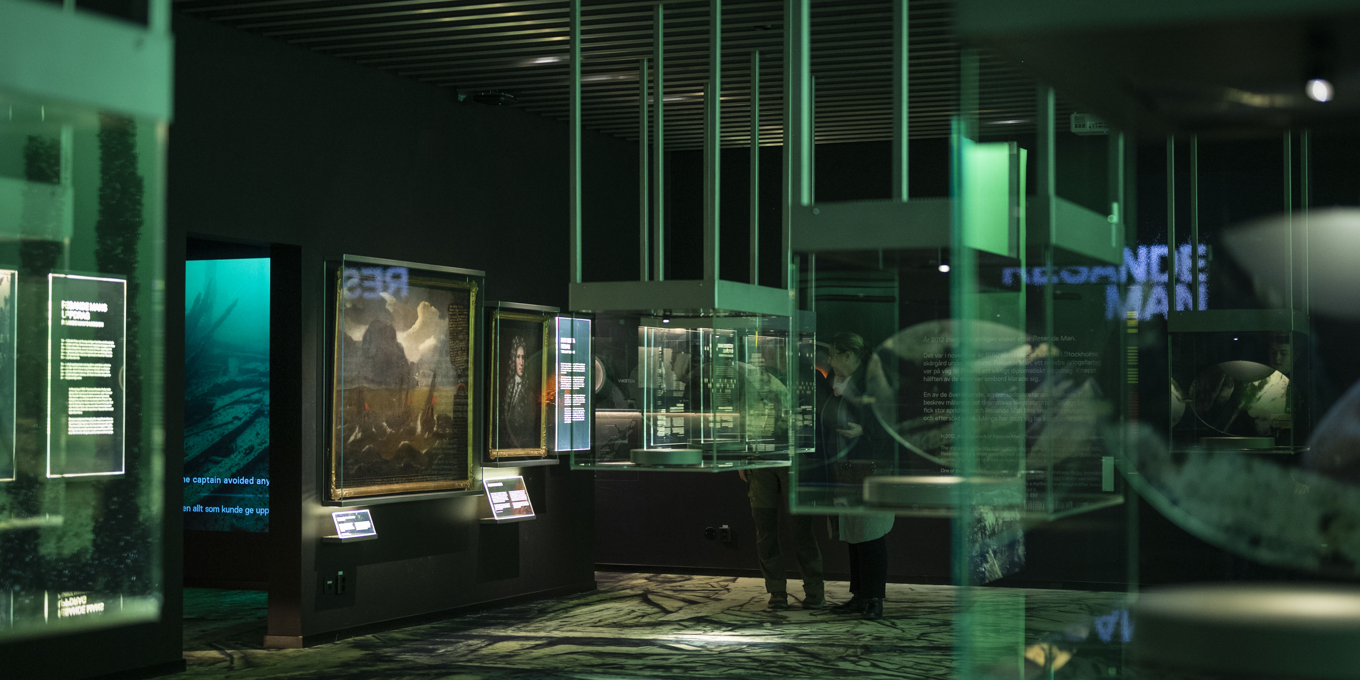 Utställningssal med montrar, målningar och digitala informationsskärmar i dämpad grön belysning.