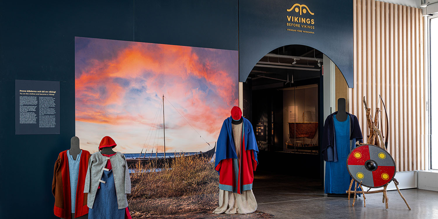 Utställning med vikingatida kläder på dockor samt en rund sköld bredvid entrén till temautställningen.