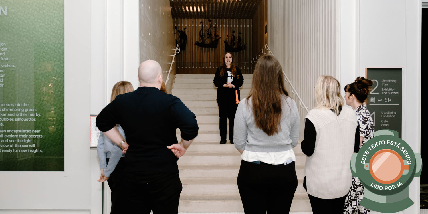 Besökare inne i museet Vraks entré tittar upp mot en trappa där en ung kvinnlig guide står, bilden är märkt med en dykhjälm och texten: Este texto está sendo lido por IA. 