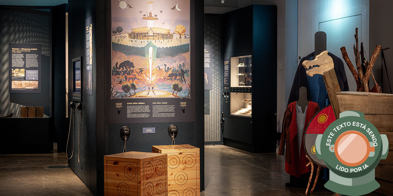 Utställningsrum med illustration av Valhall, montrar med föremål samt vikingakläder och sköld i förgrunden. Bilden är märkt med en dykhjälm och texten: Este texto está sendo lido por IA.