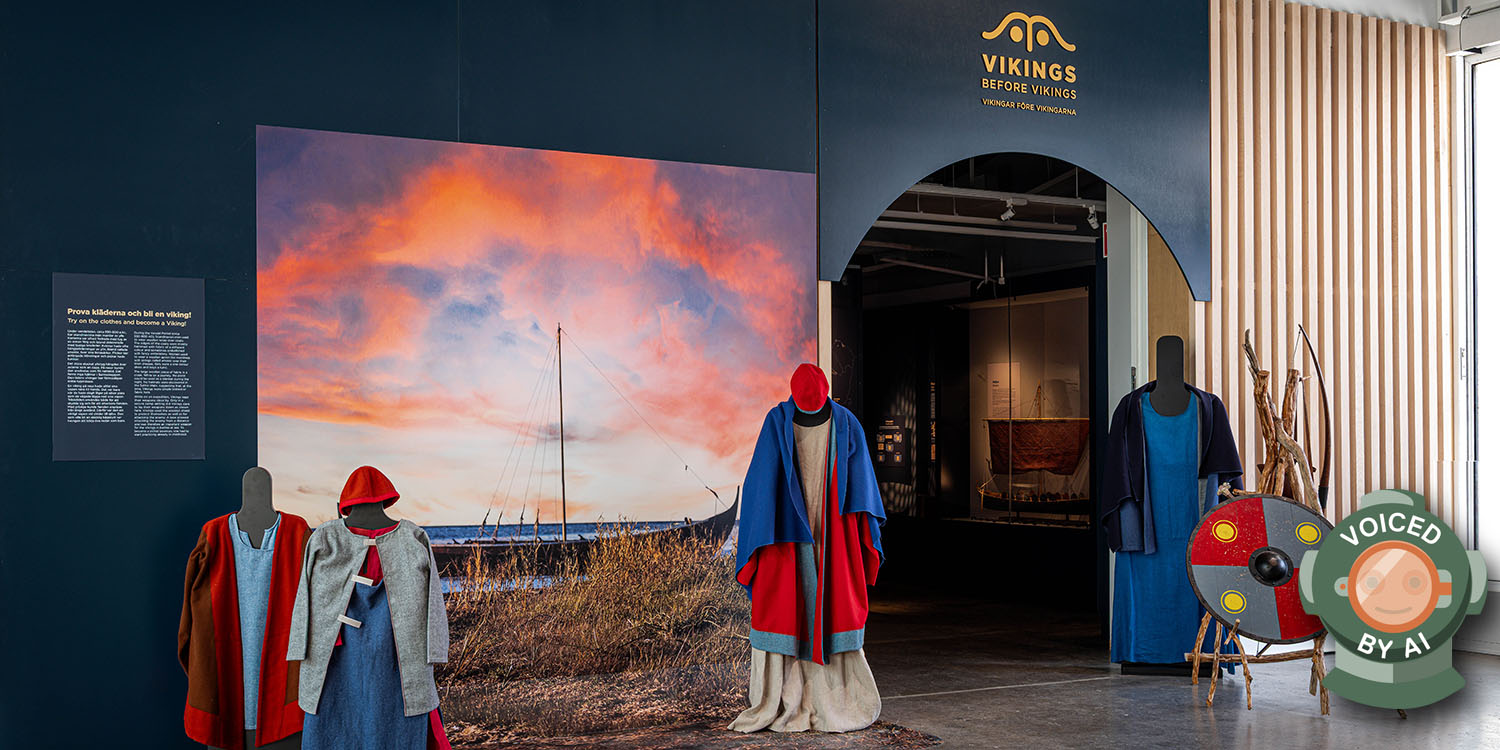 Utställning med vikingatida kläder på dockor samt en rund sköld bredvid entrén till temautställningen. Bilden är märkt med en dykhjälm och texten Voiced by AI.