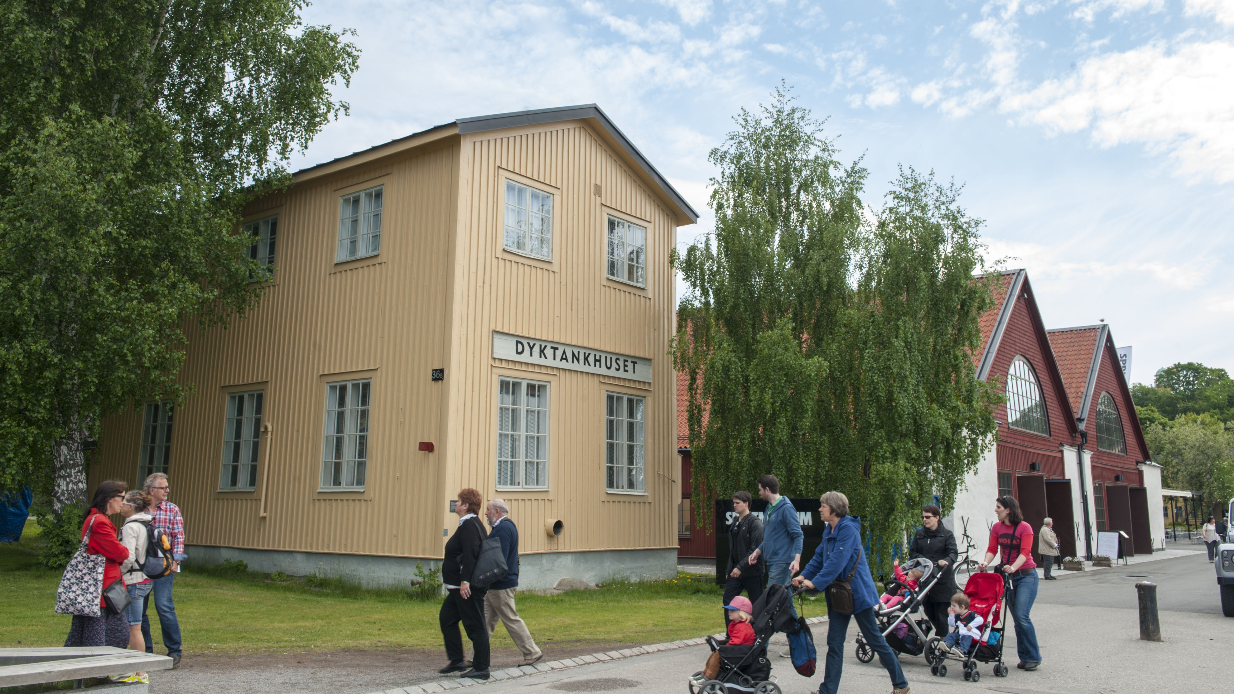 Folk promenerar förbi den ljusgula träbyggnaden Dyktankhuset på Djurgården, omgiven av träd och intill röda museibyggnader.