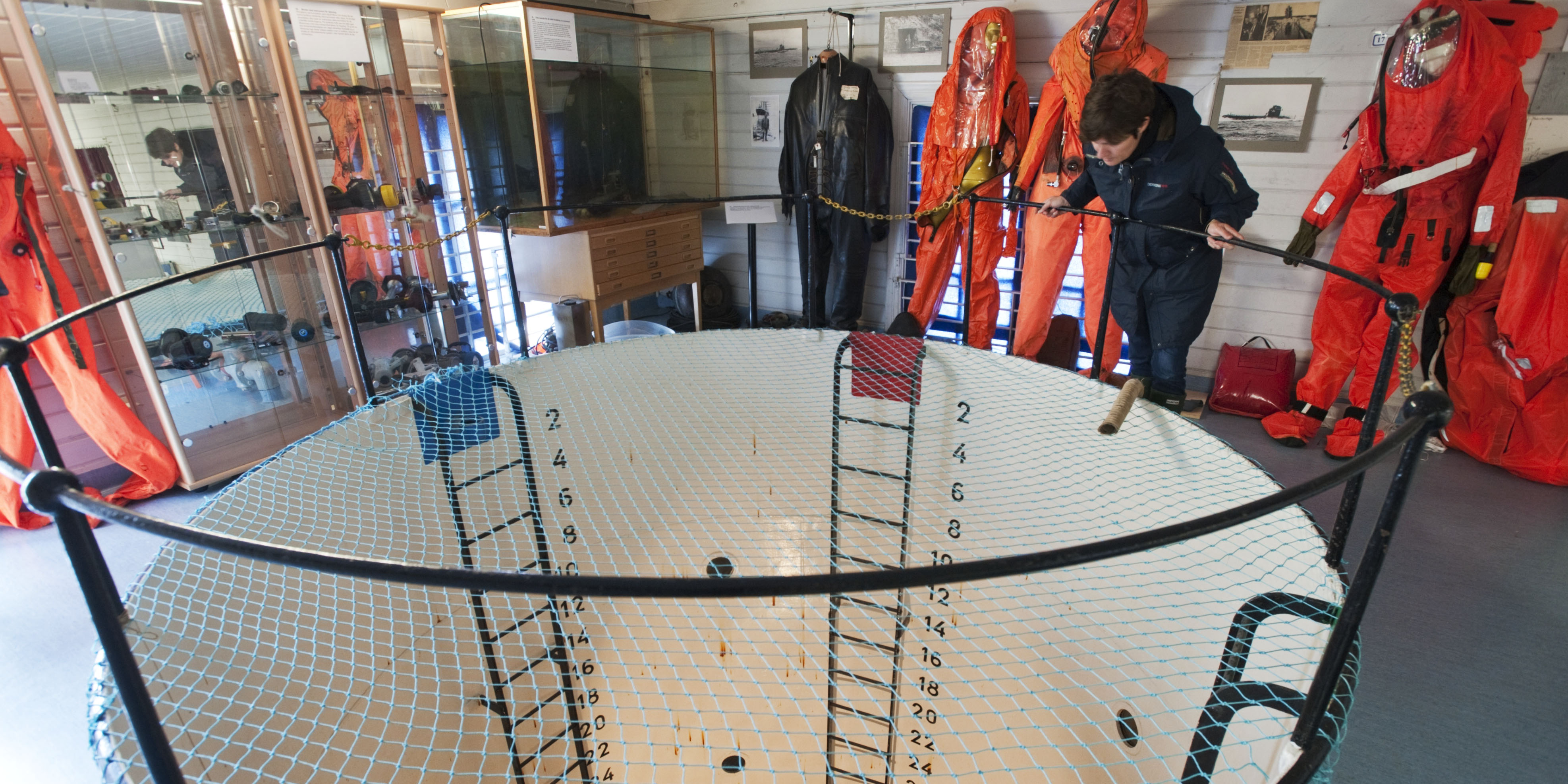 En person lutar sig över räcket runt den stora vita dyktanken inomhus, omgiven av montrar med dykutrustning och upphängda dykdräkter.