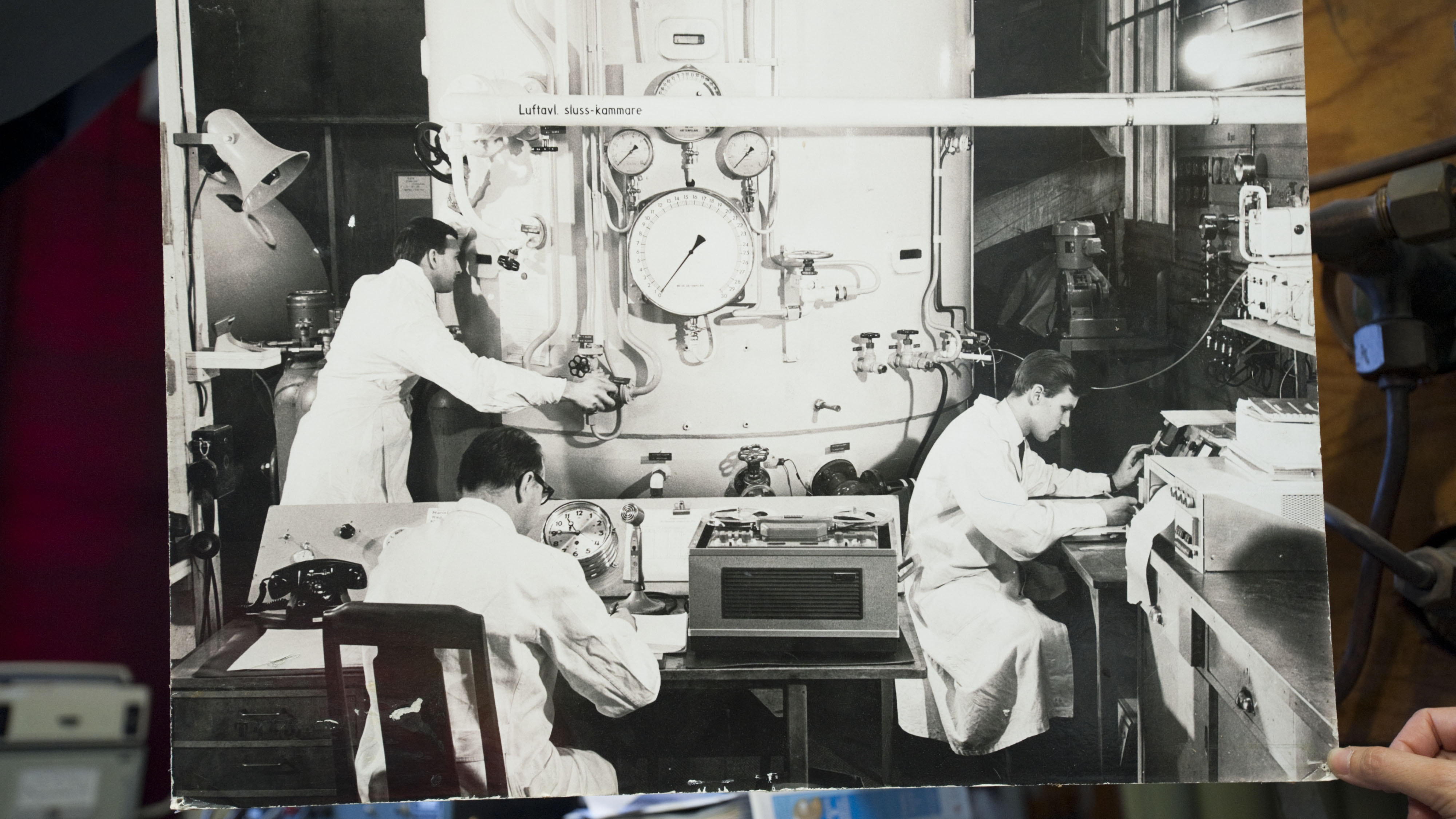 Svartvitt historiskt foto som visar tre tekniker i labbrockar som arbetar vid kontrollpaneler och instrument kopplade till dyktankens tryckkammare.