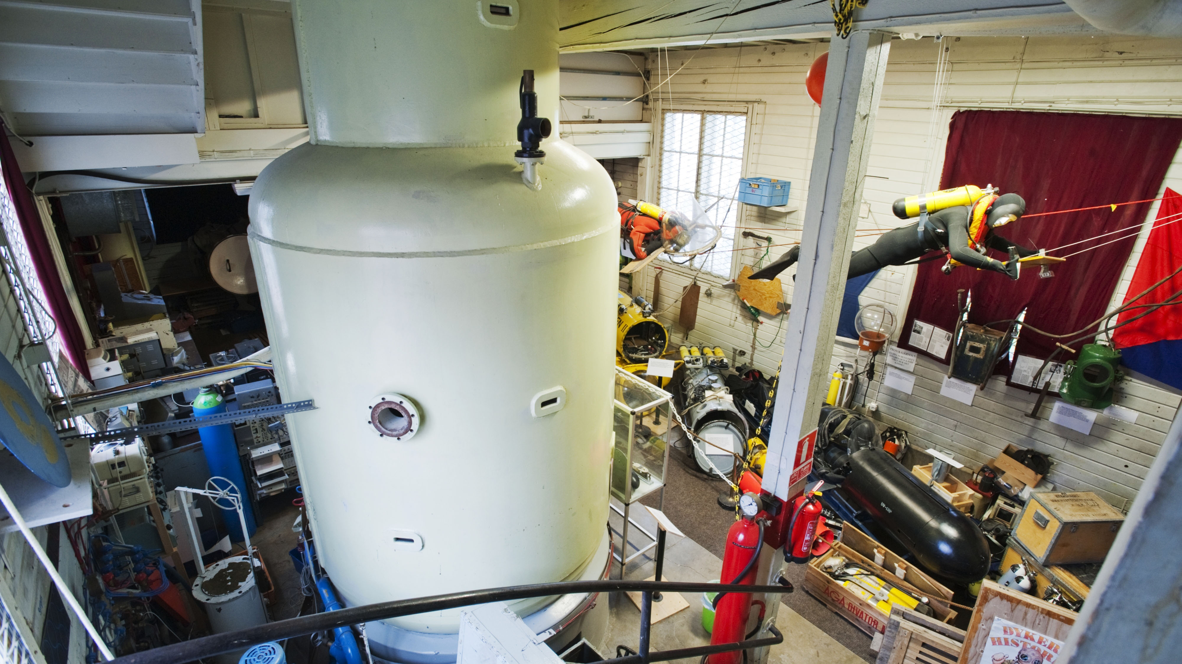 Vy över dyktankens stora cylindriska trycktank inomhus, omgiven av dykutrustning, modeller av dykare i sele och olika tekniska föremål
