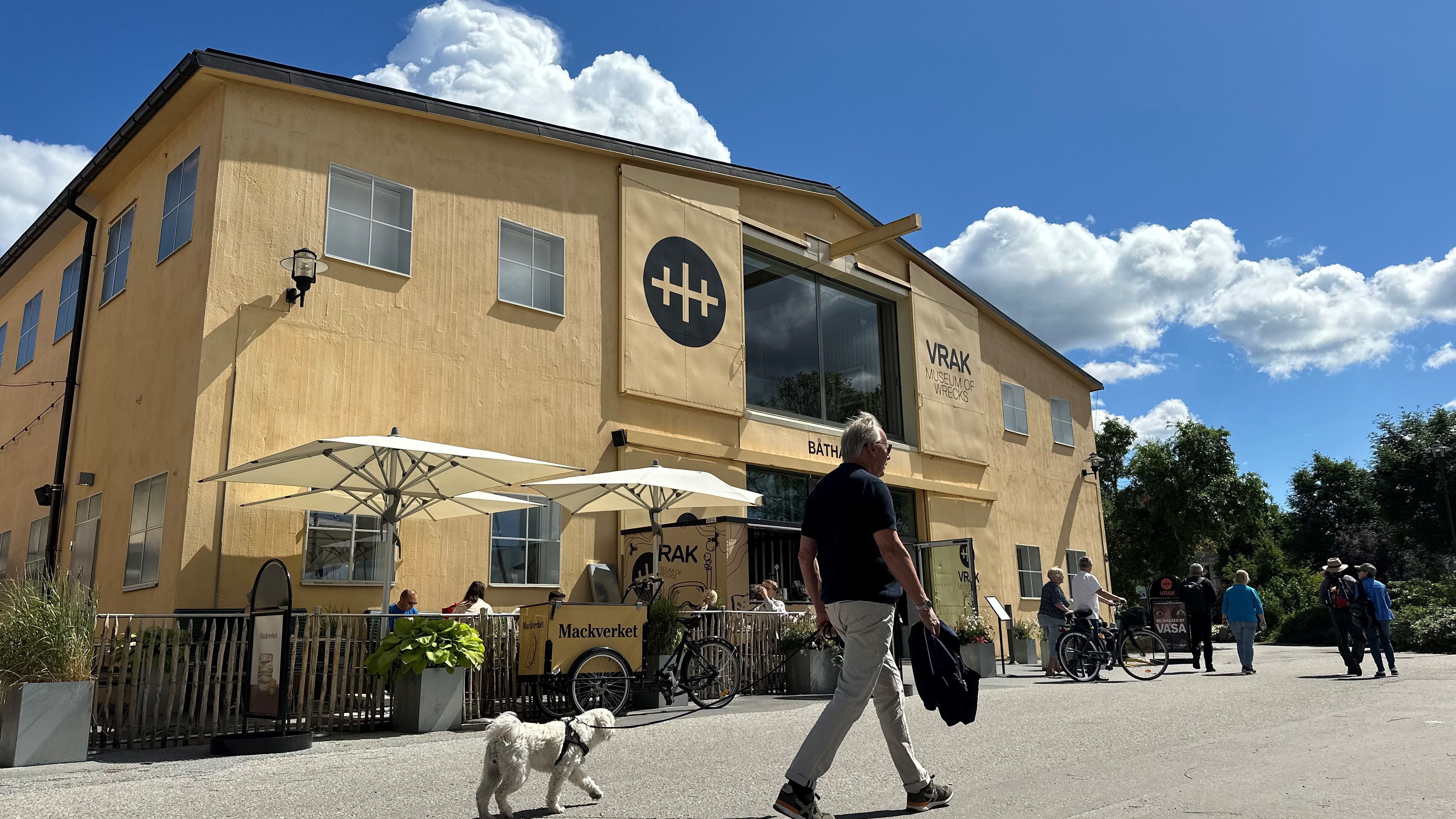 Foto som visar museet Vrak utifrån en sommardag med blå himmel. Byggnaden är ljusgul, man med hund går förbi.