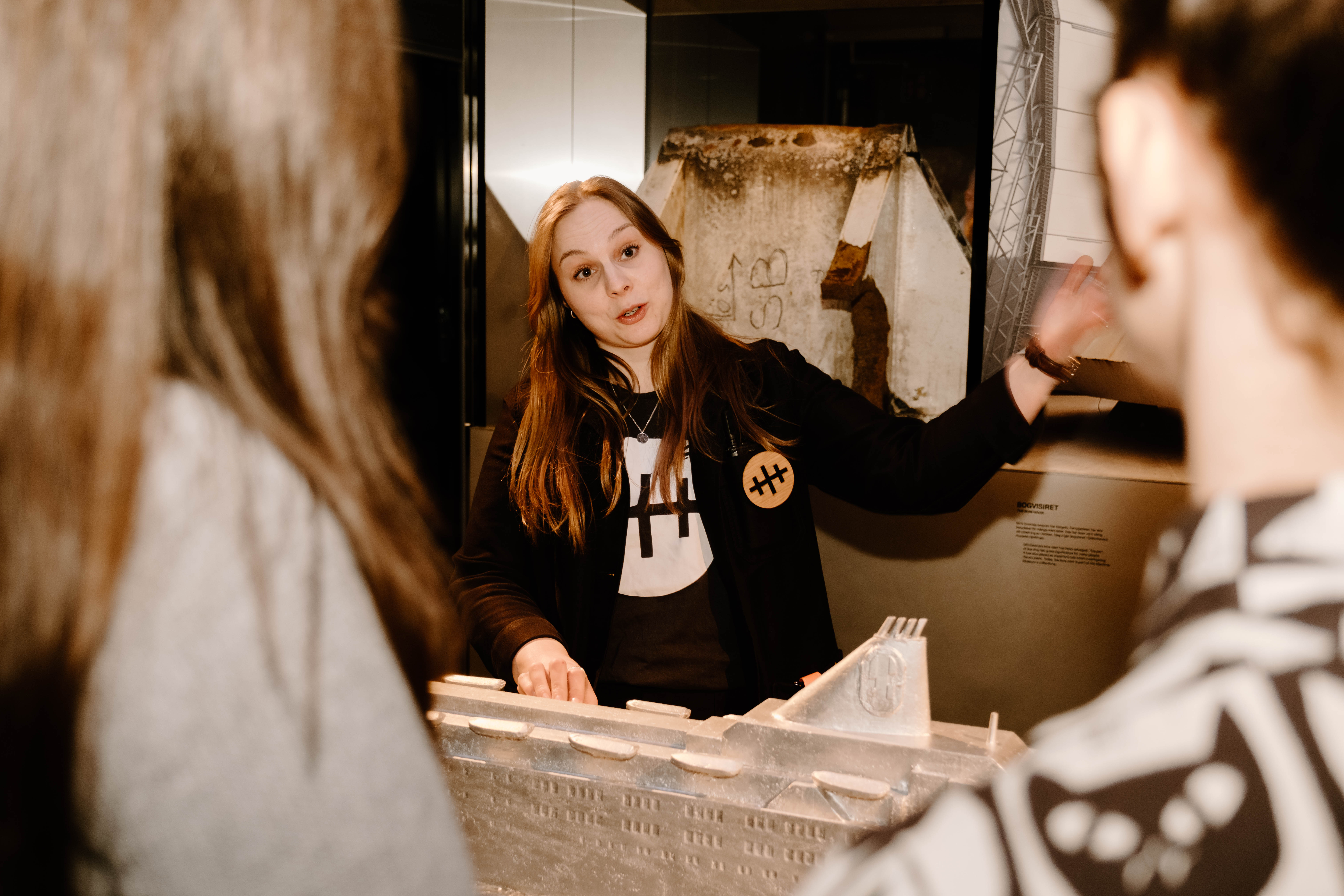A female guide shows a model of a ship, two visitors with their backs to the camera are listening.