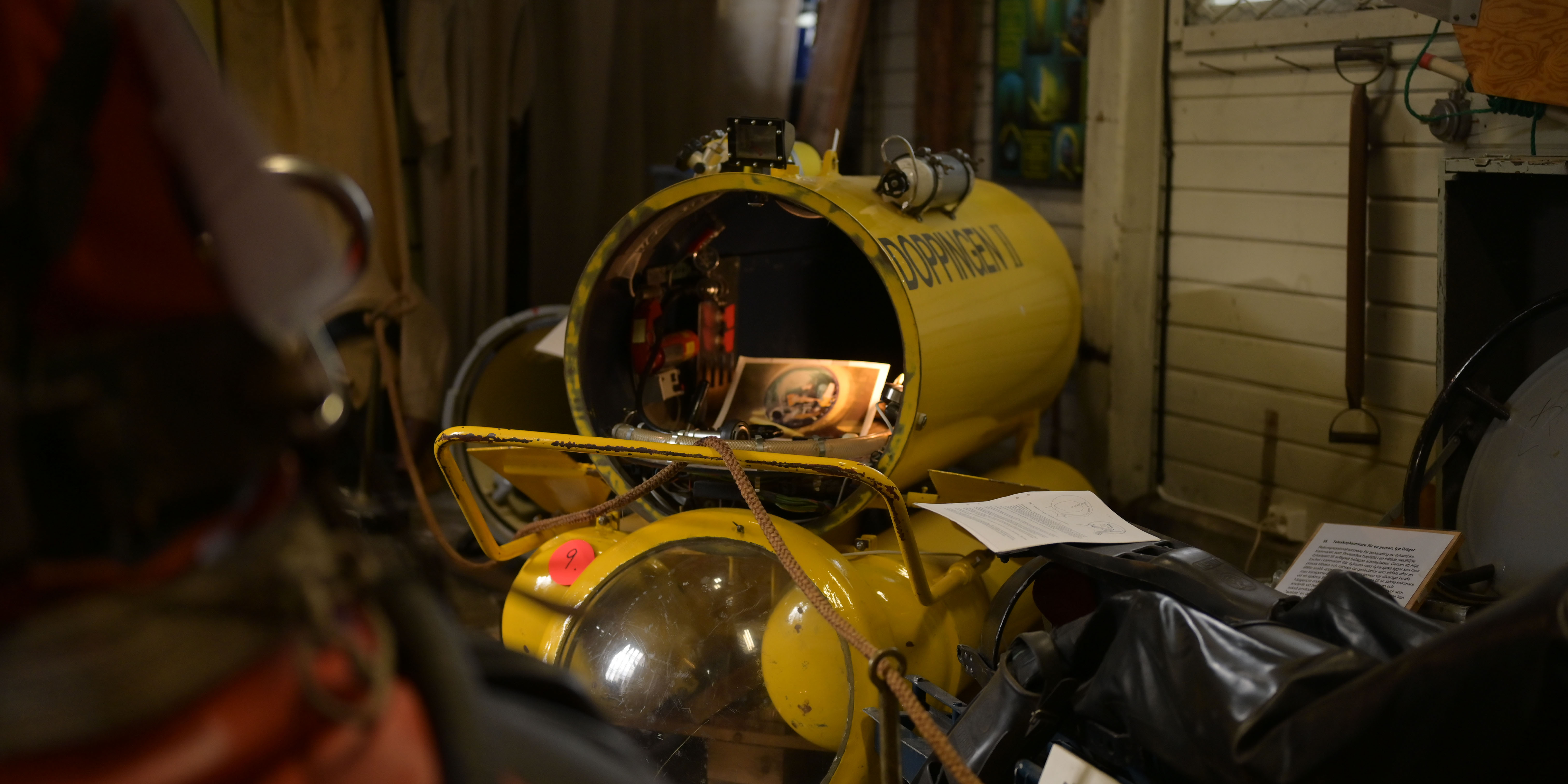 En gul miniubåt med texten ”Doppingen II” står i ett trångt förrådsutrymme. Luckan är öppen och visar instrument och kablar inuti. Runtom finns dykarutrustning och gamla föremål.