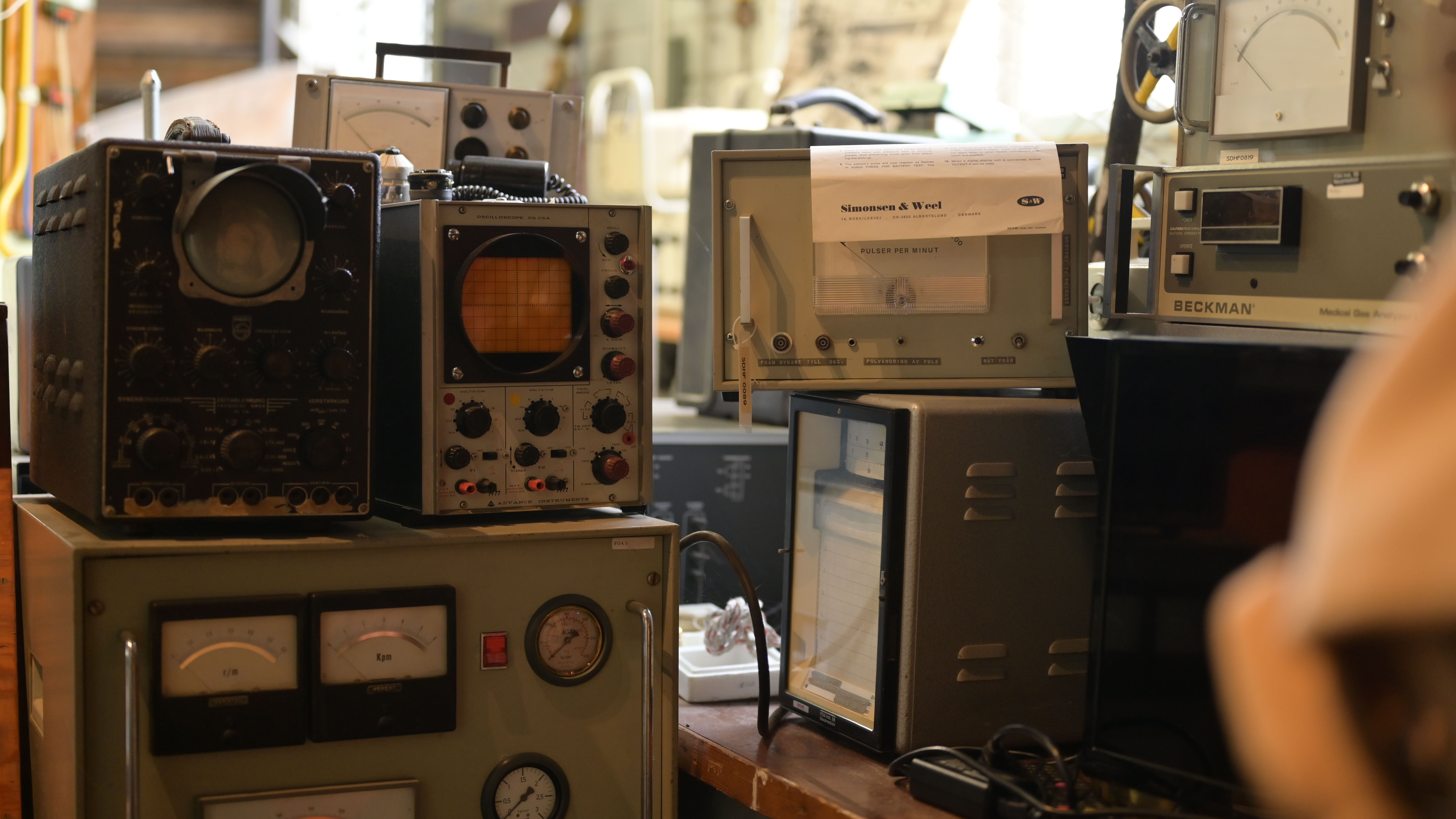 Ett bord fullt av gamla mätinstrument, oscilloskop och analysutrustning i metallhöljen. Bland märkena syns Beckman och Simonsen & Weel. Miljön påminner om ett tekniskt laboratorium eller museum.