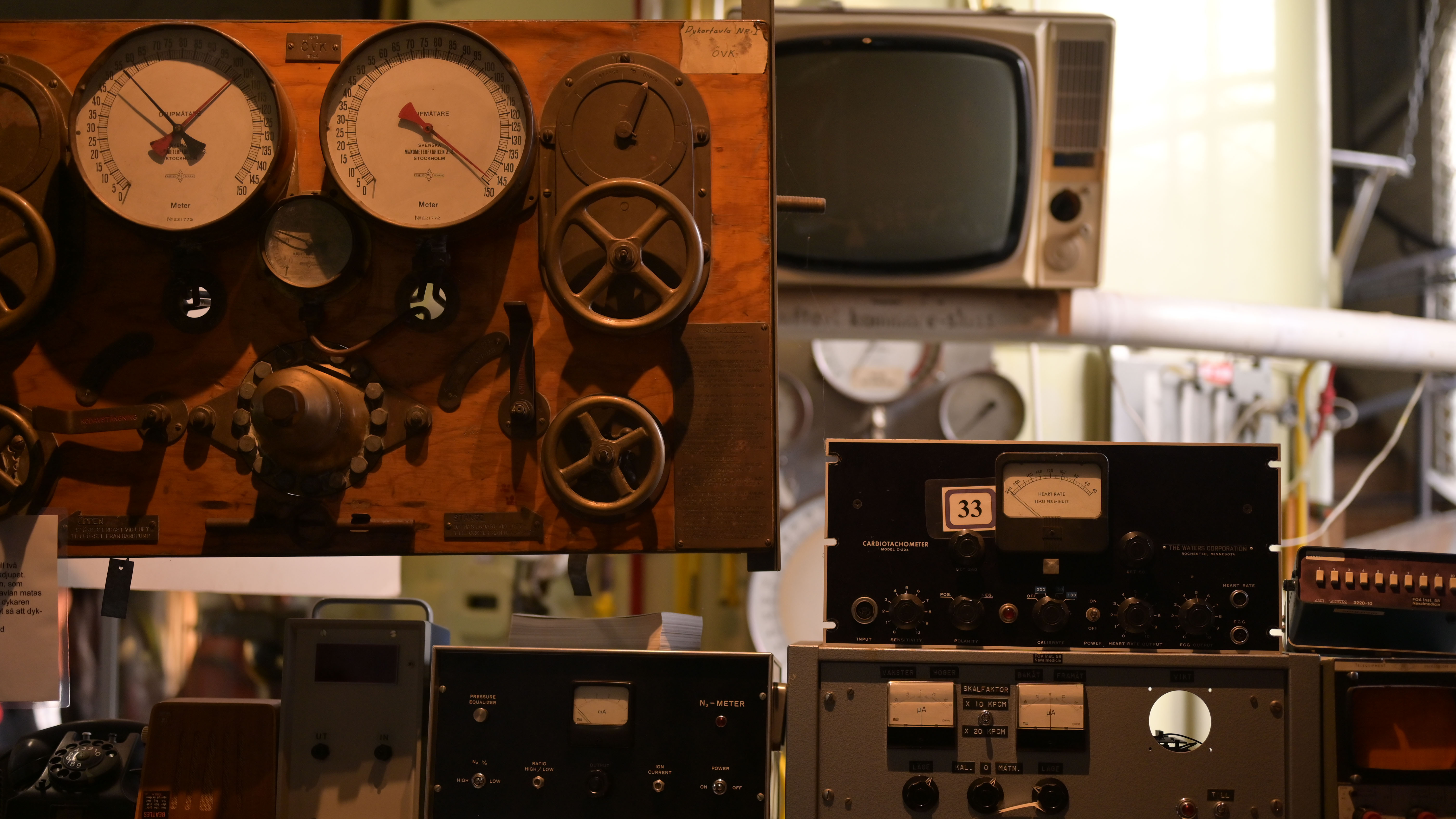 Gamla mätare och reglage monterade på en träpanel, med en äldre tv-apparat i bakgrunden. Framför står fler analoga mätinstrument och laboratorieutrustning.