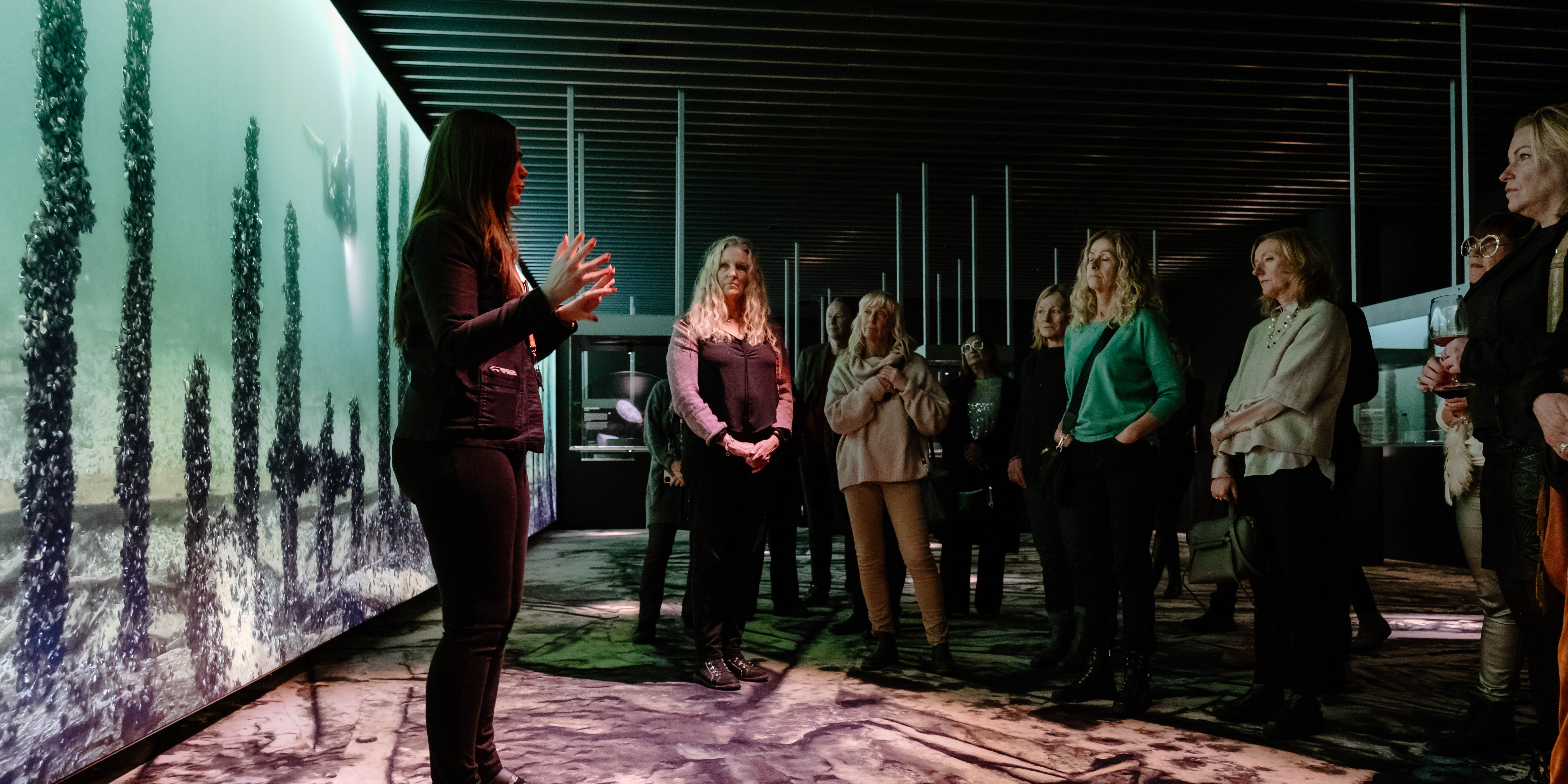 Grupp med museibesökare lyssnar på kvinnlig guide i gröntonat rum med fotoväggar. 