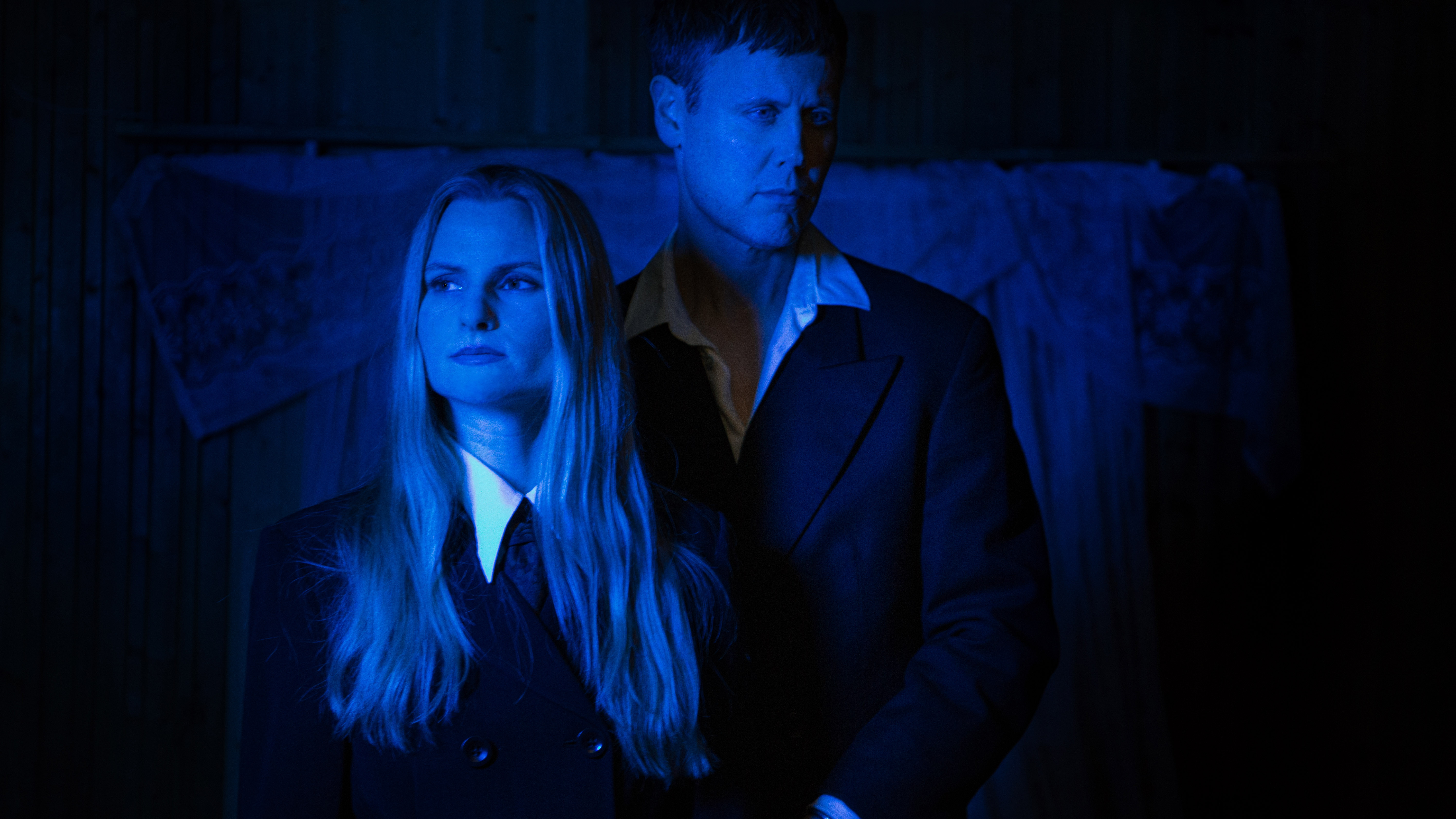 Bild i blått, svart och vitt av två personer: man  som står bakom blond kvinna båda är kläda i skjorta och kavaj. 