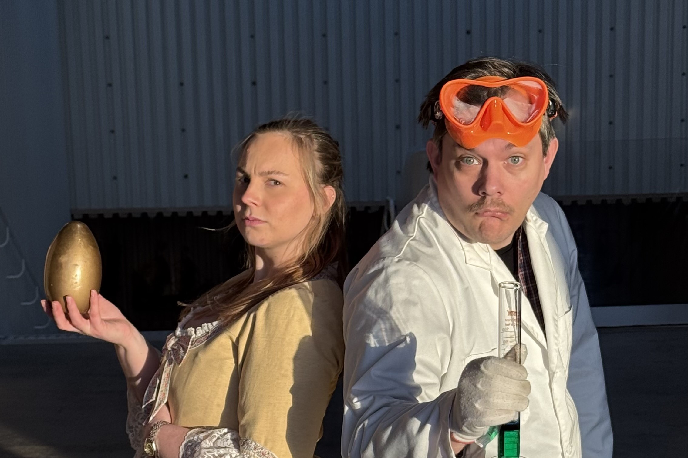 Två personer står rygg mot rygg i dramatiskt ljus. Den ena håller ett guldfärgat ägg i handen, medan den andra bär en vit labbrock, skyddsglasögon på huvudet och håller ett provrör med grön vätsk