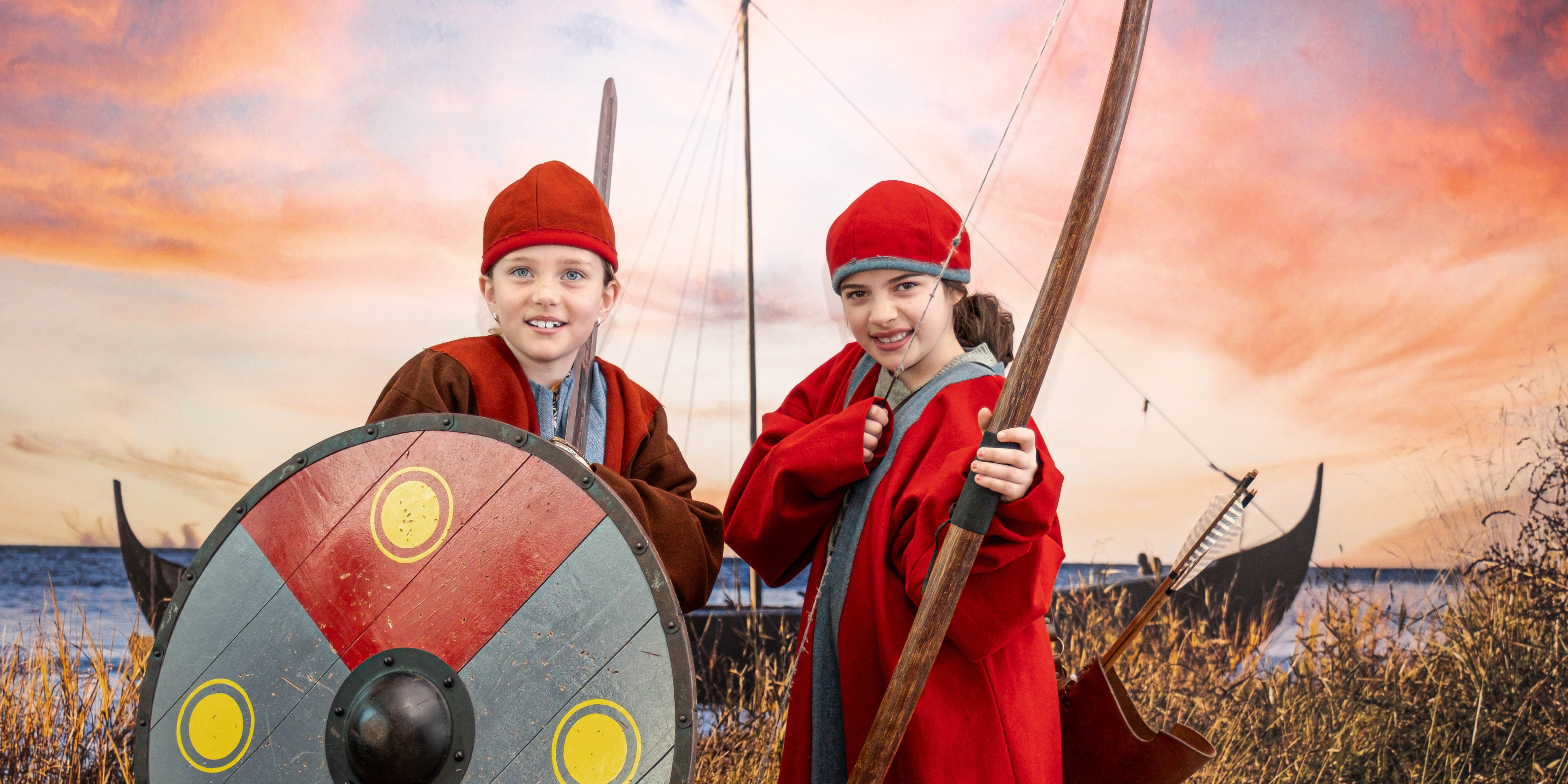 Två flickor provar vikingakläder och pilbåge. 