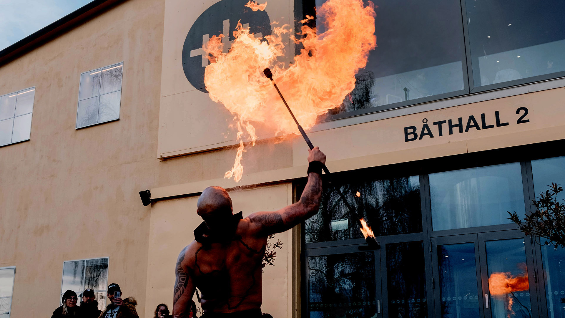 Person med rakat huvud sedd bakifrån blåser ett moln av eld utanför entrén till museet Vrak. 