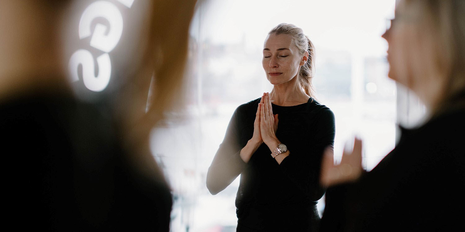 Kvinna i svarta kläder håller ihop händerna till hälsning och blundar. Bakom henne syns stort fönster mot havet, andra personer i förgrunden. 