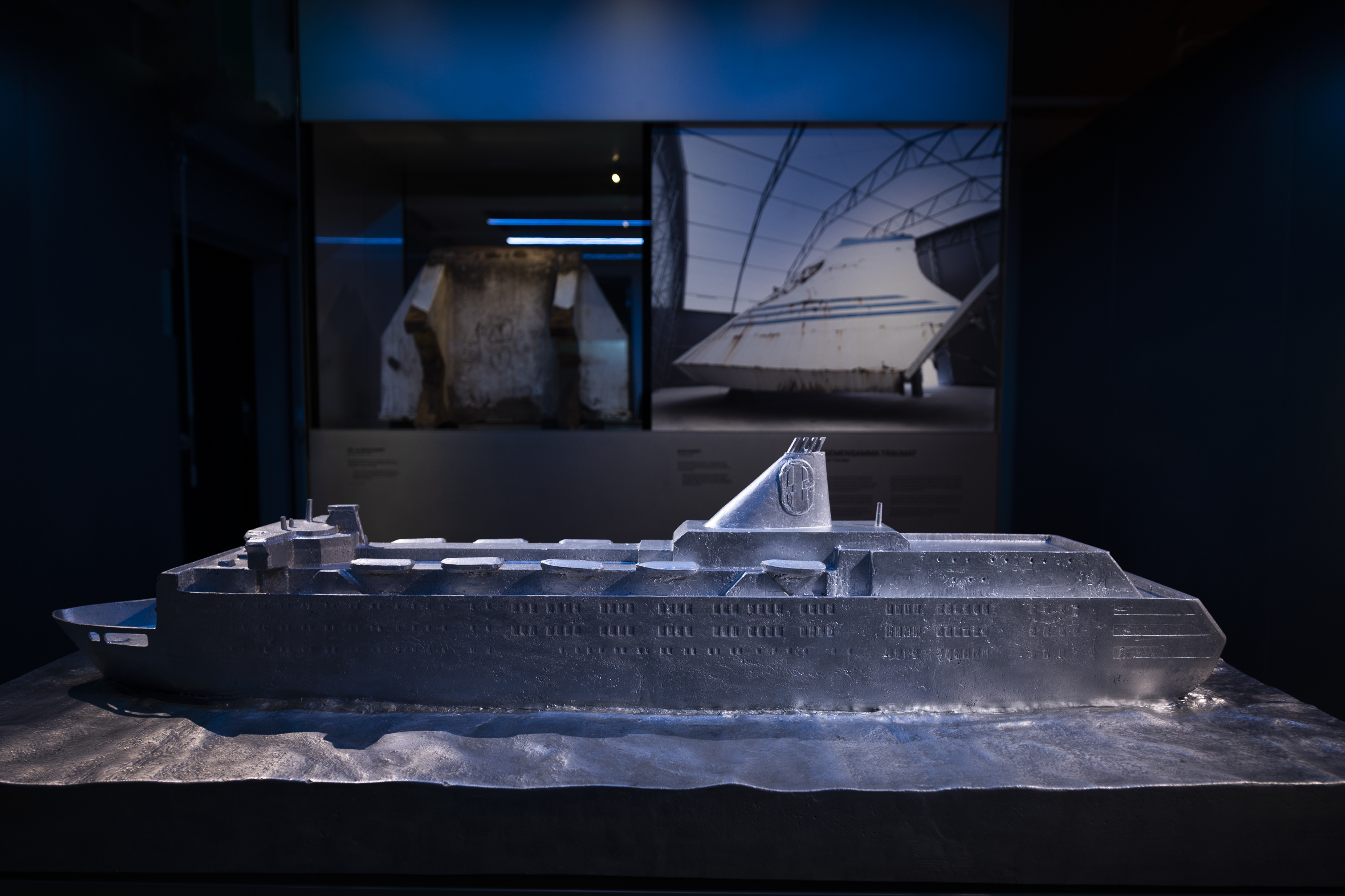 Modell av passagerarfärjan M/S Estonia i grått material visas i en mörk utställningssal, med fartygsdelar i bakgrunden.