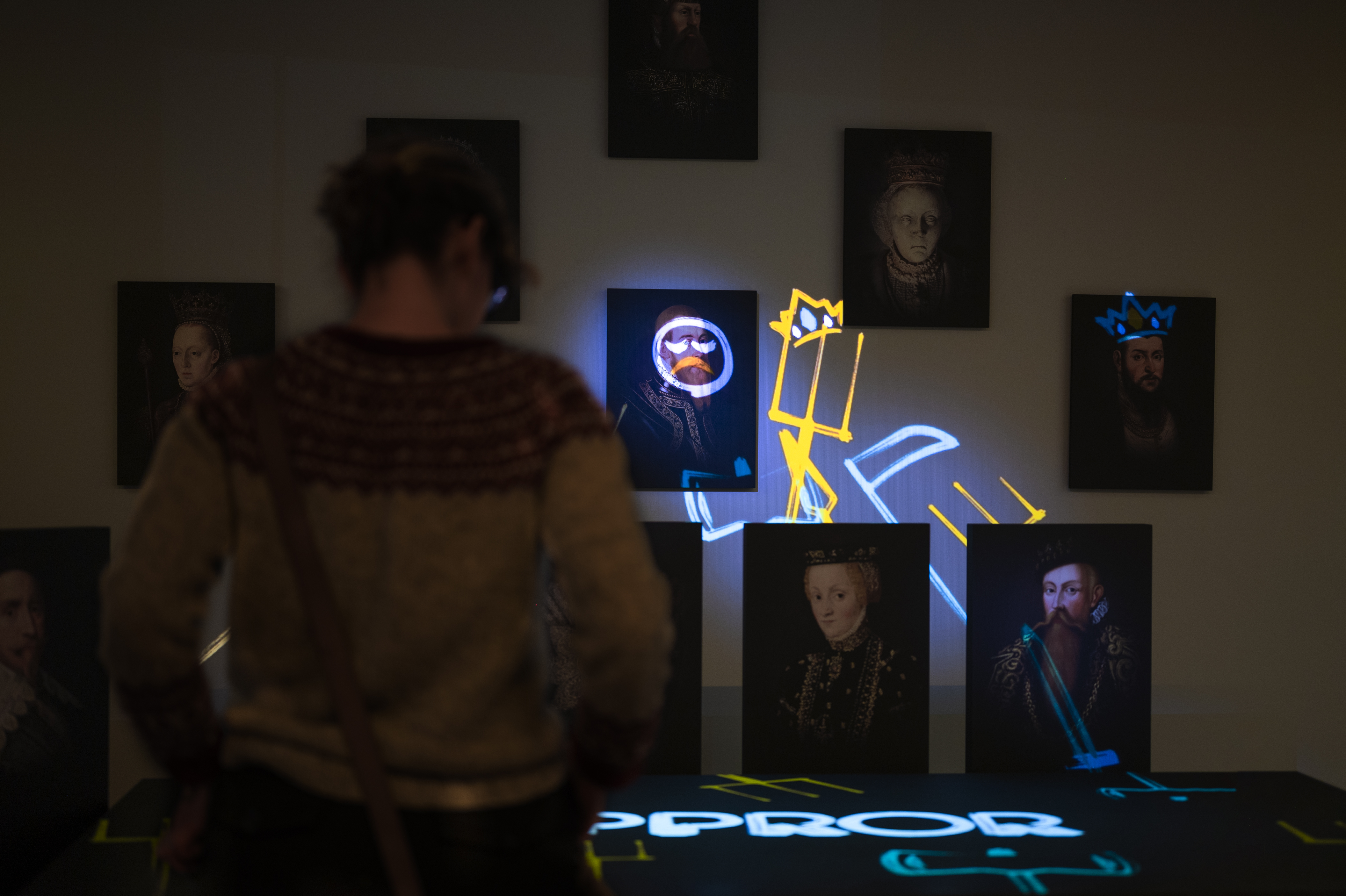 Svenska: Person i mörk utställningssal betraktar porträtt av kungligheter upplysta med färgade ljusprojektioner i blått och gult.