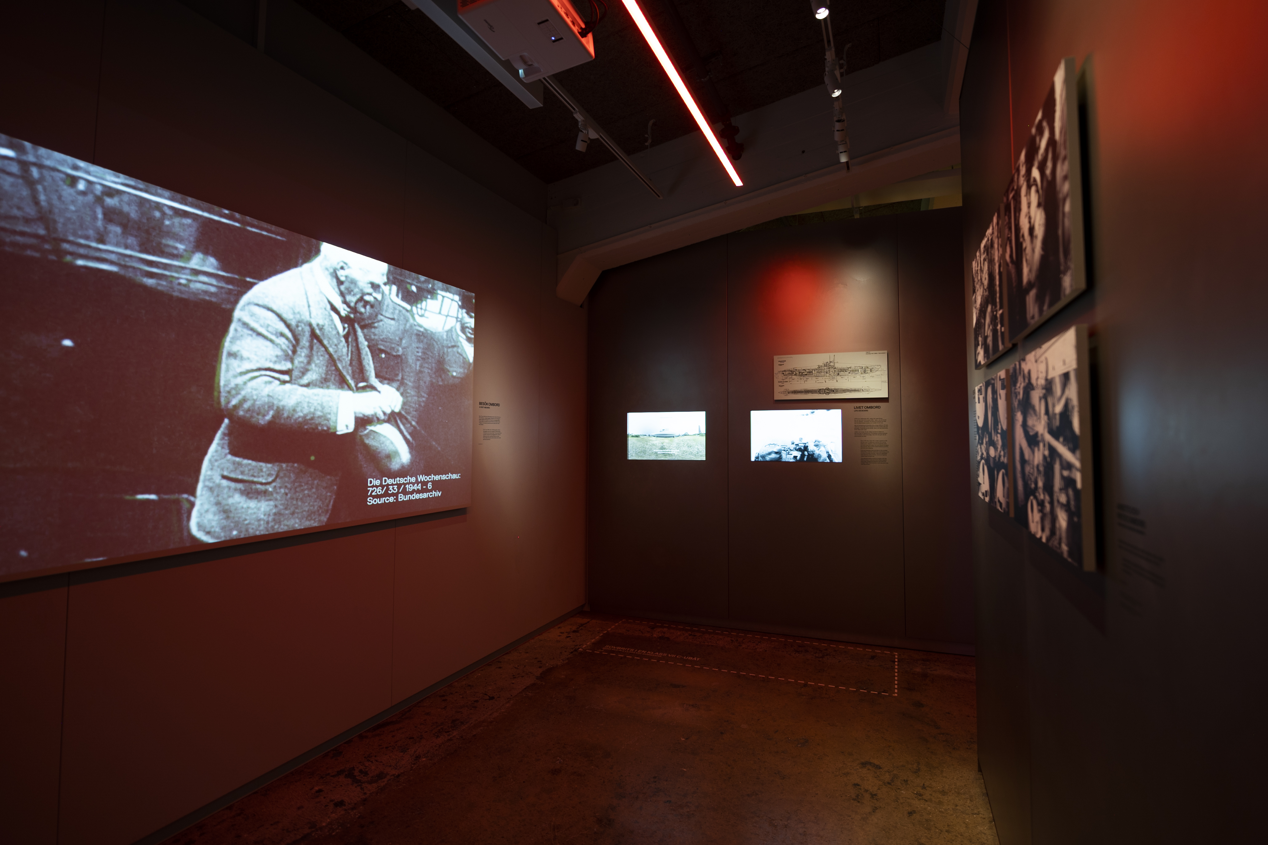  Mörk utställningssal med historiska filmklipp och fotografier projicerade på väggar med rött ljus.