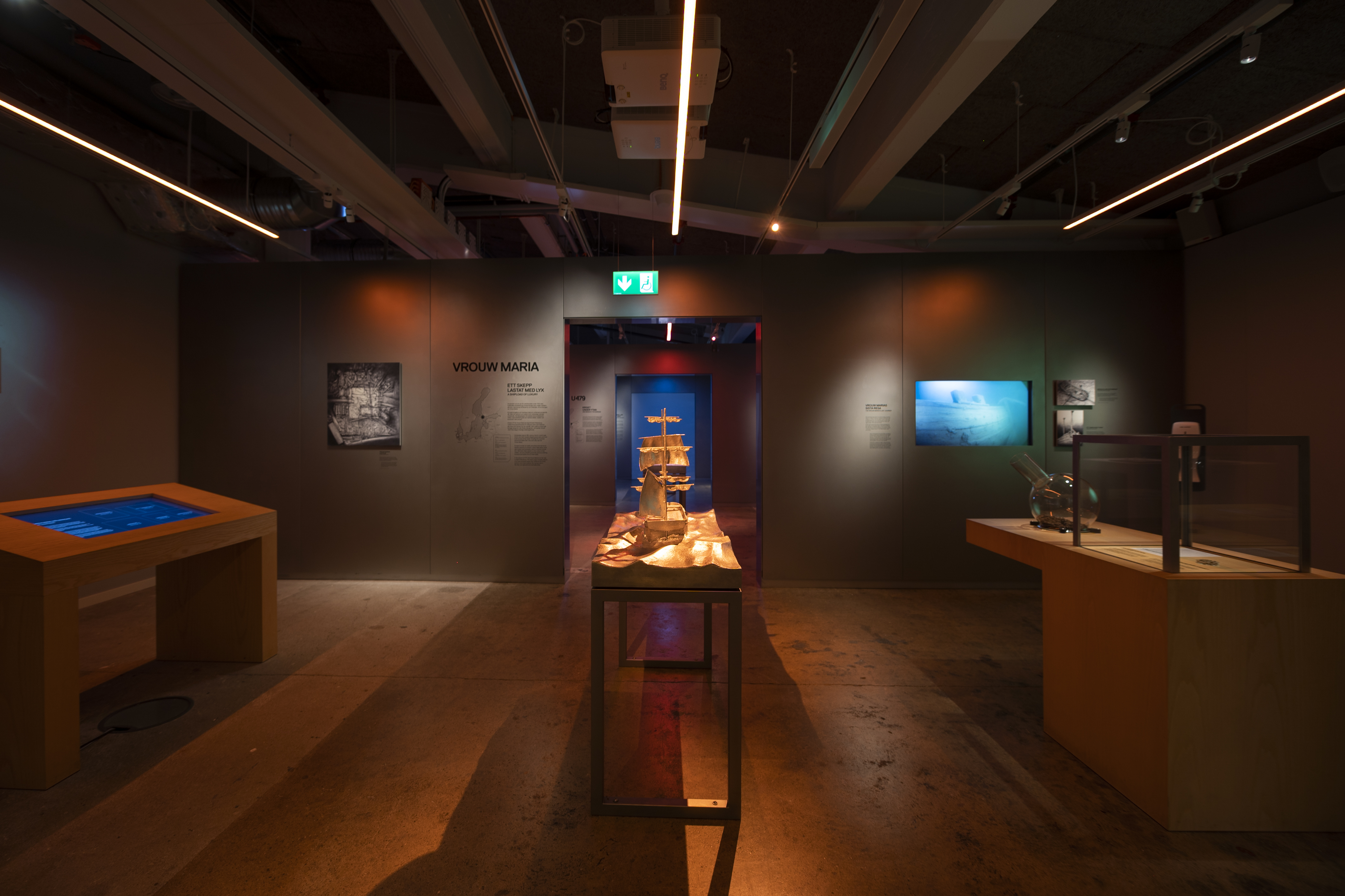 Engelska: Exhibition room with a model of the sailing ship Vrouw Maria in the center, surrounded by screens and information panels under warm lighting.
