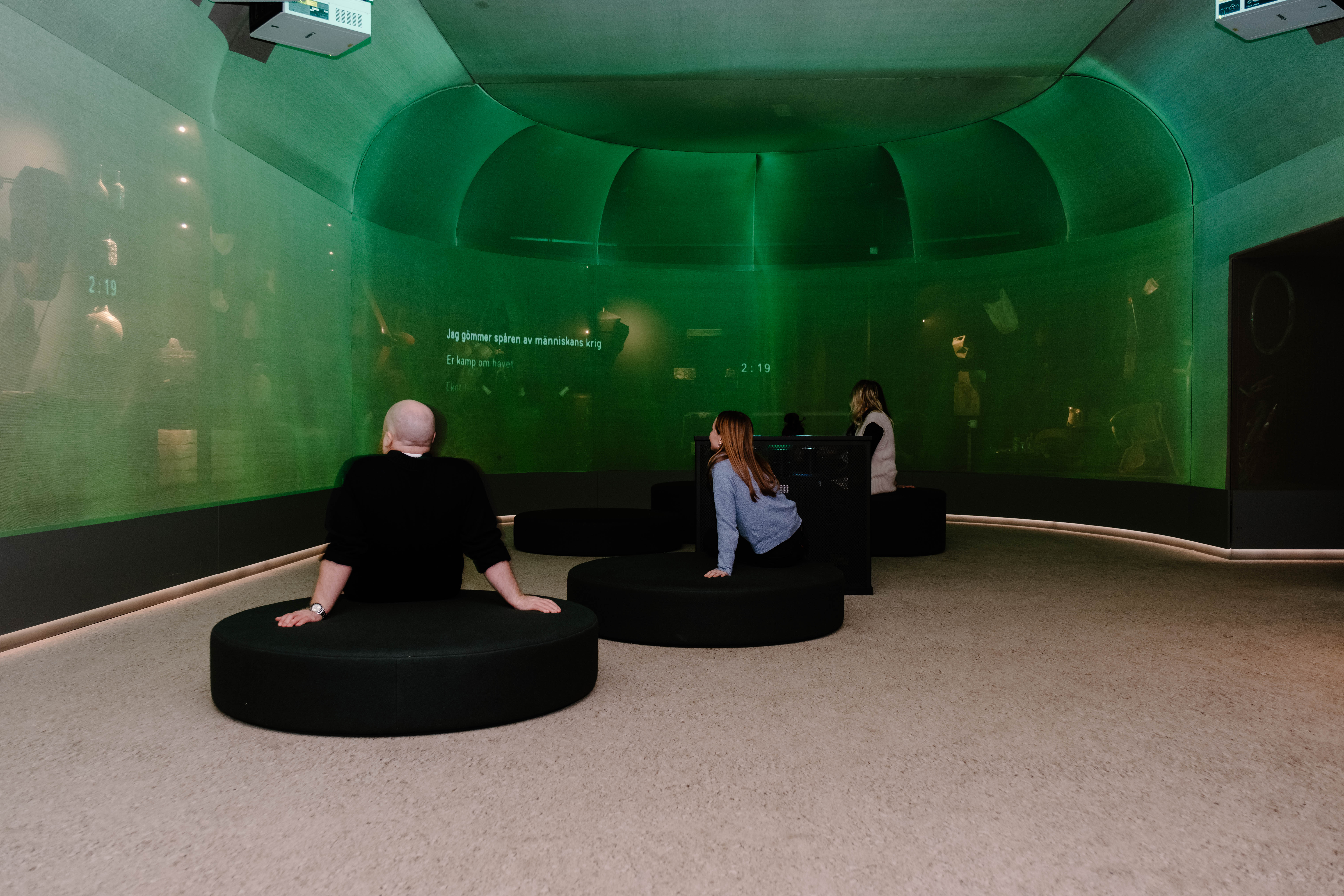 Gröntonad filmduk täcker ett välvt rum, tre personer sitter på runda soffor på golvet och tittar på projiceringen. 