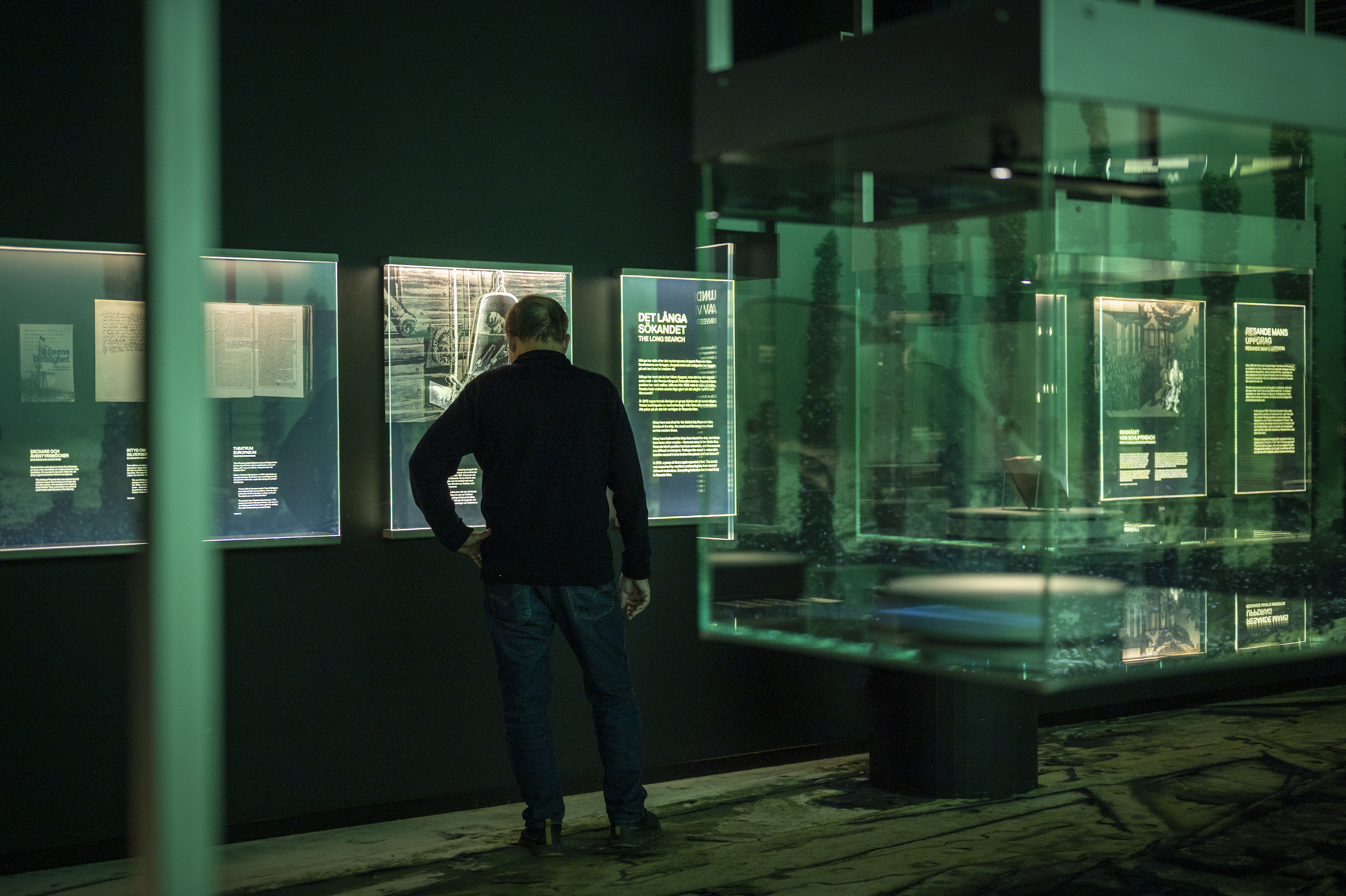 A man stands in a dimly lit museum, reading illuminated information panels with texts and images behind glass. The exhibition has a green glow and a modern design with floating glass displays.