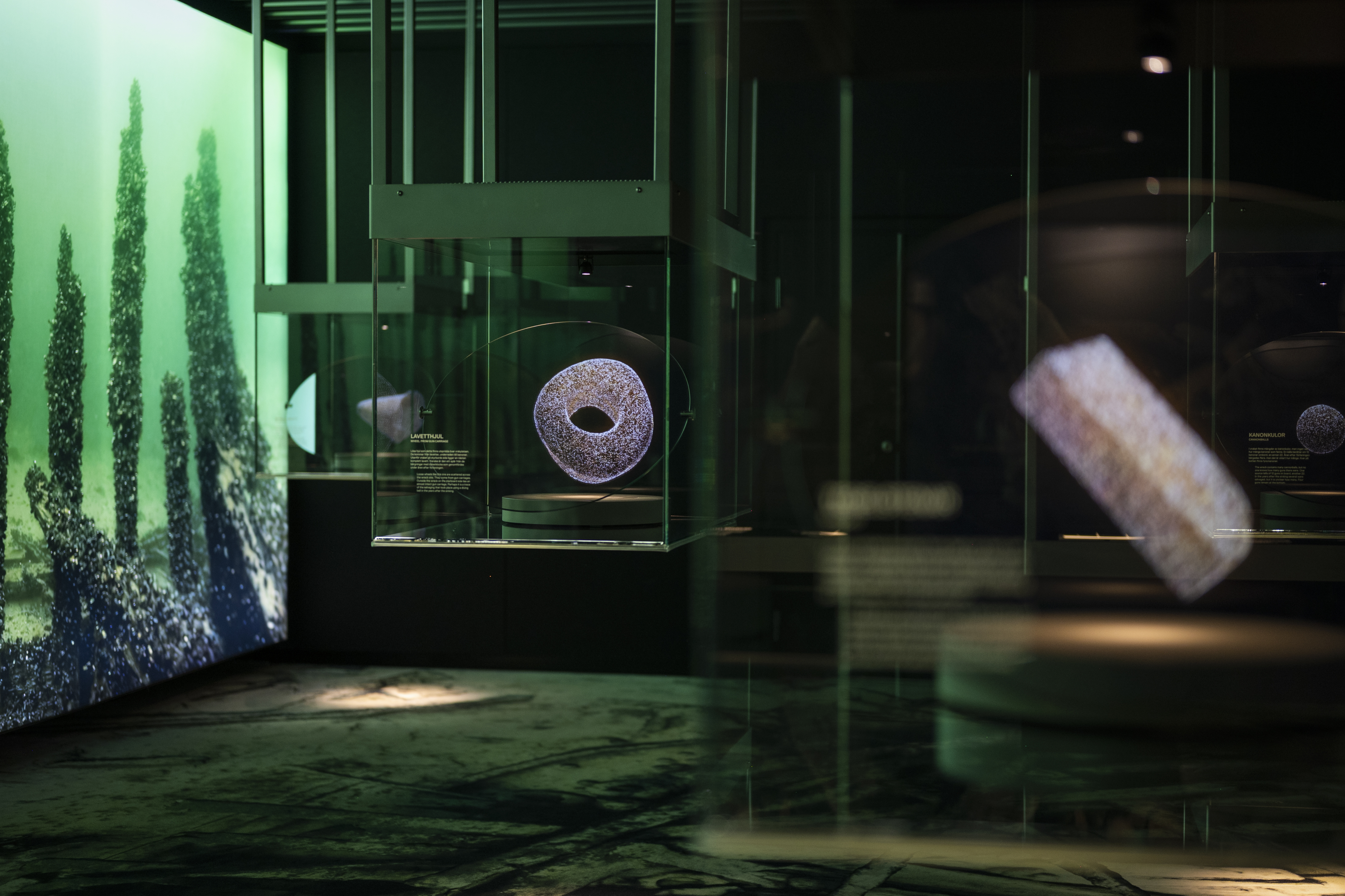 Närbild av en glasmonter i ett museum med ett runt föremål i centrum, upplyst i blått och grönt ljus. Bakom syns fler montrar och en vägg med undervattensmotiv.