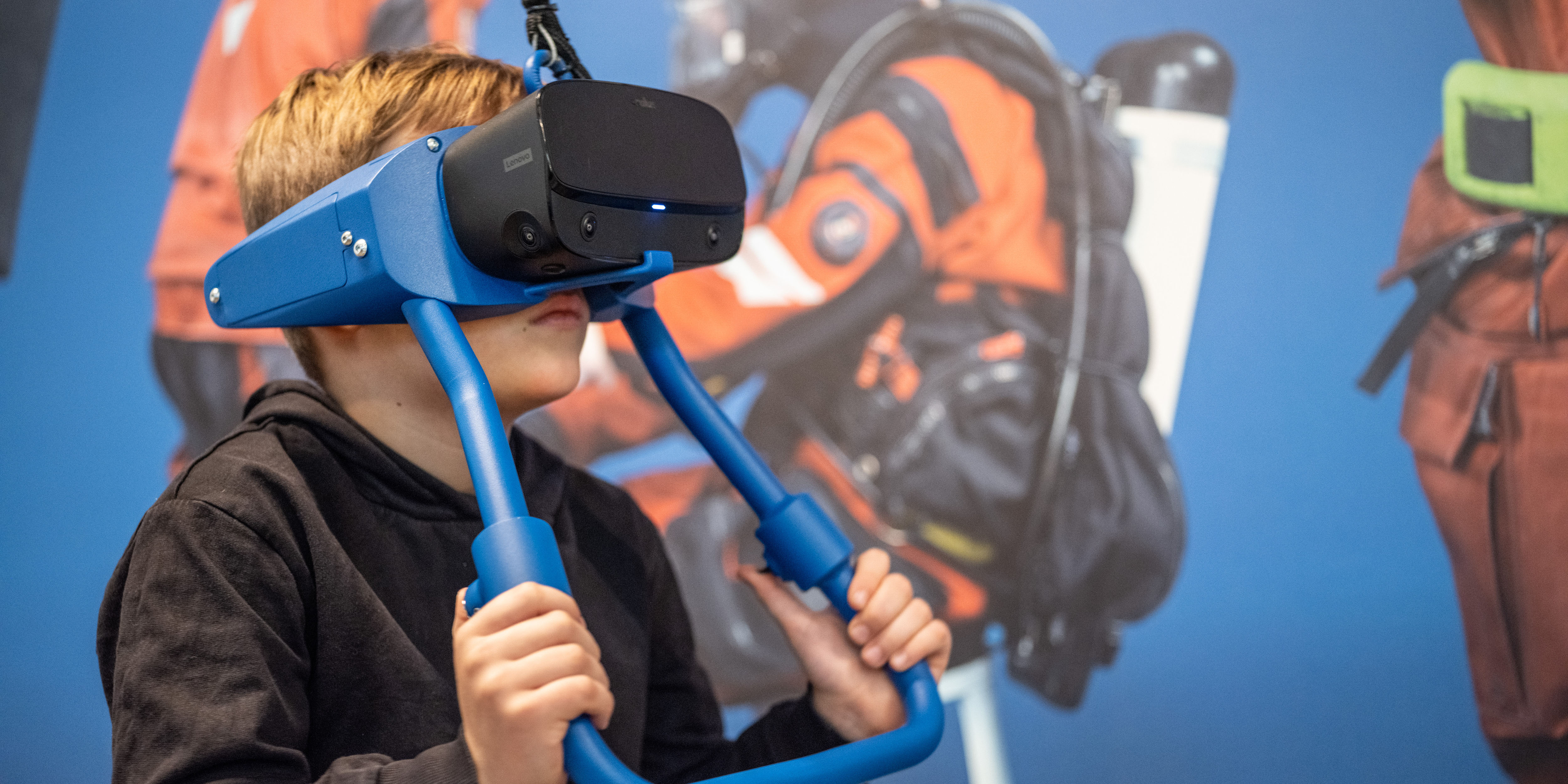 Alt-text (svenska): Ett barn använder ett VR-headset med blå ram i en utställning med dykare på väggen.