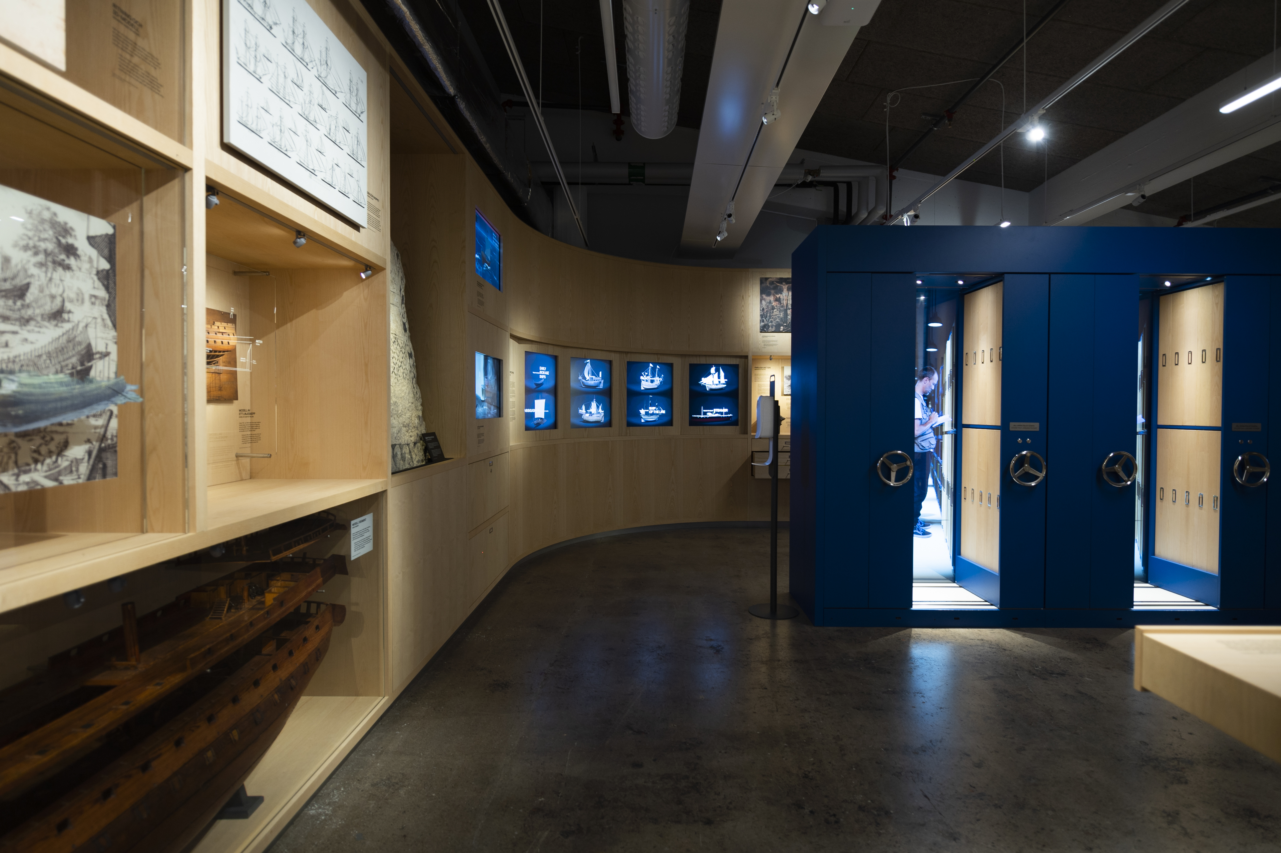 Ett museum med träpaneler, skärmar som visar fartygsmodeller och blå förvaringsskåp med rattar.