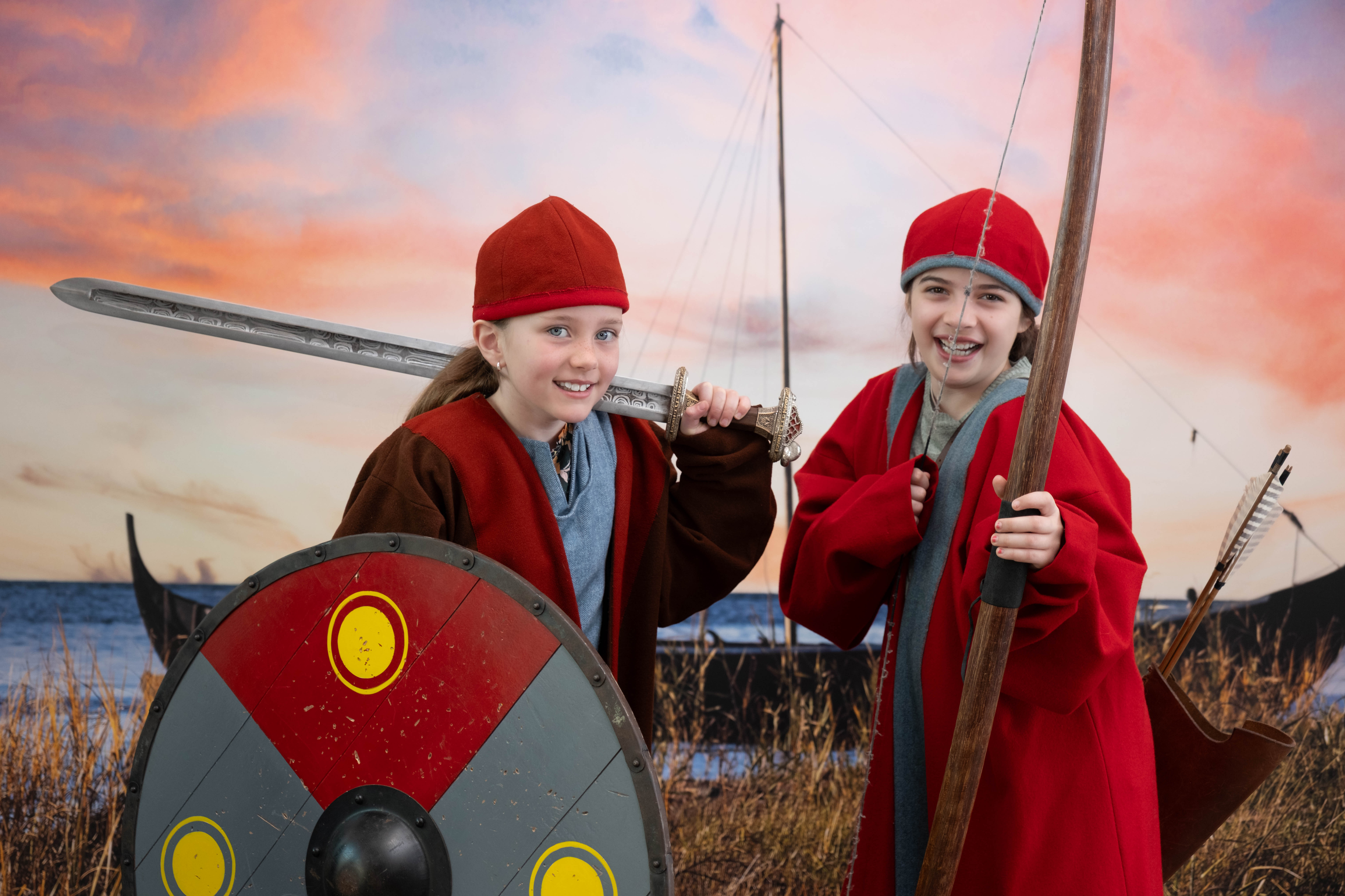 Two children dressed in Viking-inspired costumes smiling and posing with a sword, bow, and shield in front of a backdrop showing Viking ships at sunset.