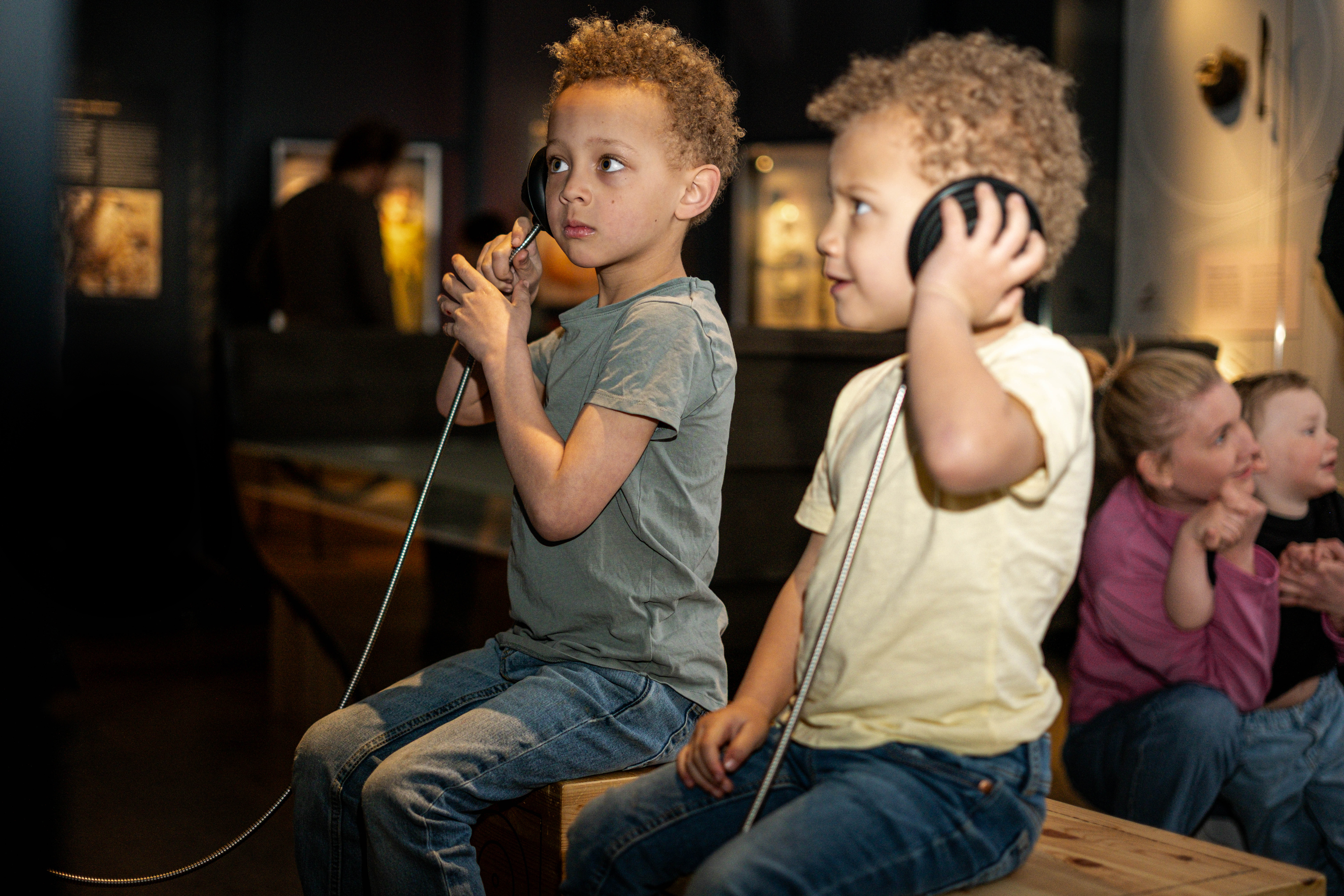 Two children sitting on wooden benches listening through headphones, with other children visible in the background in an interactive museum section.